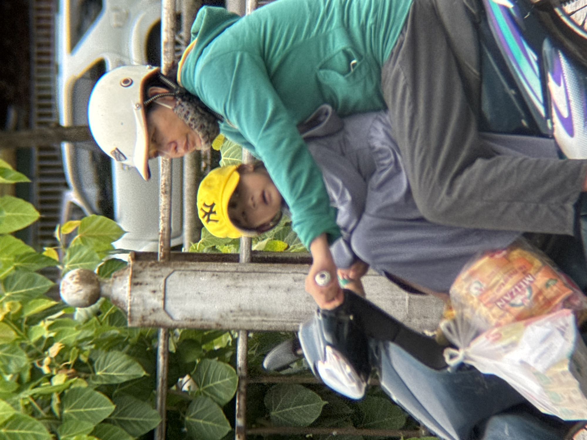 In Thuận Hóa Ward, Vietnam, a person wearing a white helmet and a teal hooded jacket is operating a dark-colored scooter. The person is seated, holding the handlebars, and is looking slightly to their left. A child, wearing a yellow cap with a black emblem, is seated in front of the person, also looking forward. A plastic bag containing items is hanging from the front of the scooter. The background shows green foliage, a metal fence, and a white vehicle. The environment appears to be outdoors, likely a street or roadside.
