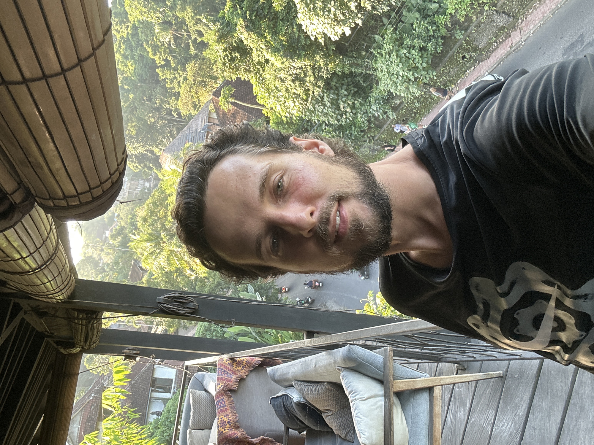 A man with a beard and dark, wavy hair is taking a selfie. He is wearing a black t-shirt with a gray graphic design. The image is taken from a high angle, looking down and to the side. In the background, lush green trees and houses can be seen. A road with several motorbikes is visible below. The lighting suggests it is either sunrise or sunset, with warm golden light filtering through the trees. The man's expression is relaxed and he is smiling slightly. In the foreground, there are some outdoor furniture pieces, including pillows and a woven rug. The overall setting appears to be an outdoor balcony or patio area, possibly in a tropical location. The context provided indicates the location is the Ubud District, Indonesia.