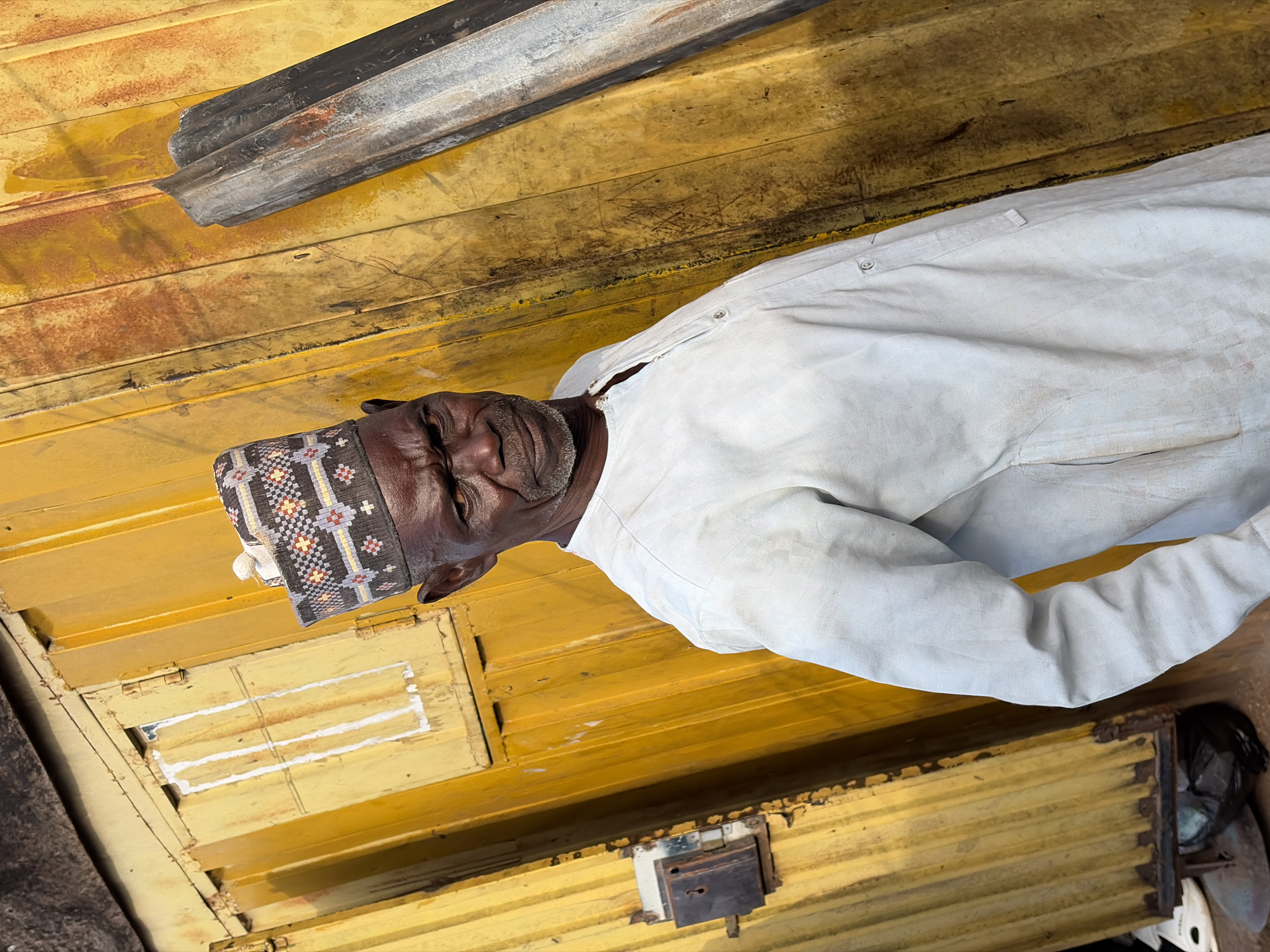 An older man, with dark skin and a short grey beard, is featured in a close-up, head-and-shoulders shot, looking directly at the camera with a calm, slightly neutral expression. He wears a plain white, long-sleeved shirt and a distinctive cap (likely a kufi) adorned with intricate black, white, red, and yellow geometric patterns.
The man is situated within what appears to be an enclosed, utilitarian space, possibly a market stall, a small room, or the interior of a vehicle, consistent with the Gura Top, Nigeria location context. The background is characterized by aged, textured yellow surfaces, resembling painted wood or corrugated metal, which show visible signs of wear, dirt, and rust. A dark, weathered metal beam runs diagonally across the upper left part of the frame. In the lower right, a metallic latch mechanism is visible on one of the yellow surfaces, suggesting a door or a storage unit.
The lighting appears to be natural daylight, casting some soft shadows. No specific activity is occurring other than the man posing for the photograph. No visible text is present in the image. The scene conveys a candid portrait captured within a humble, well-used environment.