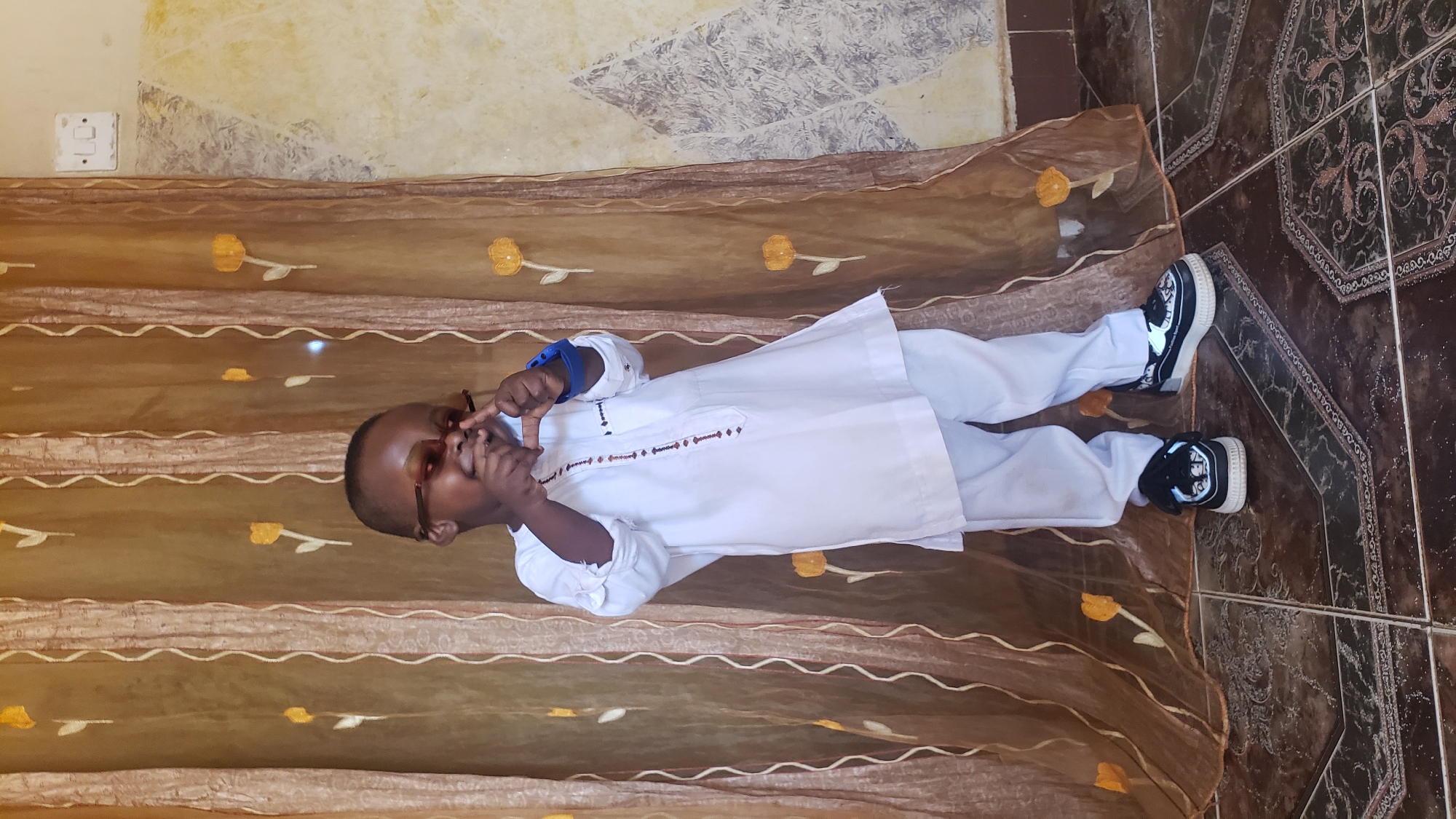 A young child wearing glasses, a white tunic, white pants, and black-and-white sneakers is lying on a brown and tan patterned cloth with yellow flower designs, on a brown tiled floor, making a framing gesture with their hands. The location is the city of Narayi, Nigeria. The background includes a wall with patchy paint above the cloth.