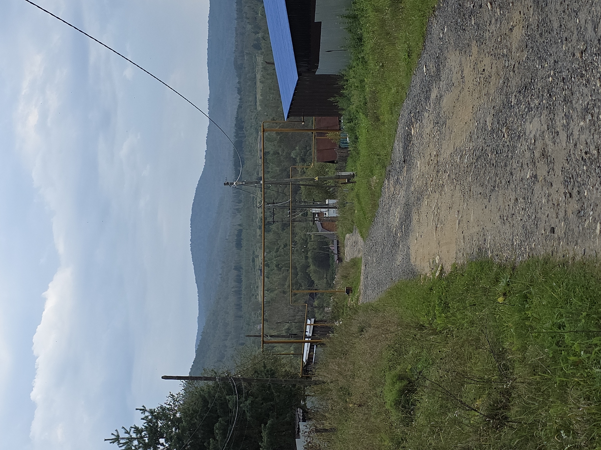 A dirt road leads through a rural settlement with low-rise buildings and dense vegetation. The road is lined with grass and weeds on either side. In the background, rolling hills covered in trees recede into the distance under a cloudy sky. Yellow pipes, likely utility lines, run horizontally across the scene. The overall impression is one of a remote, somewhat undeveloped area.
