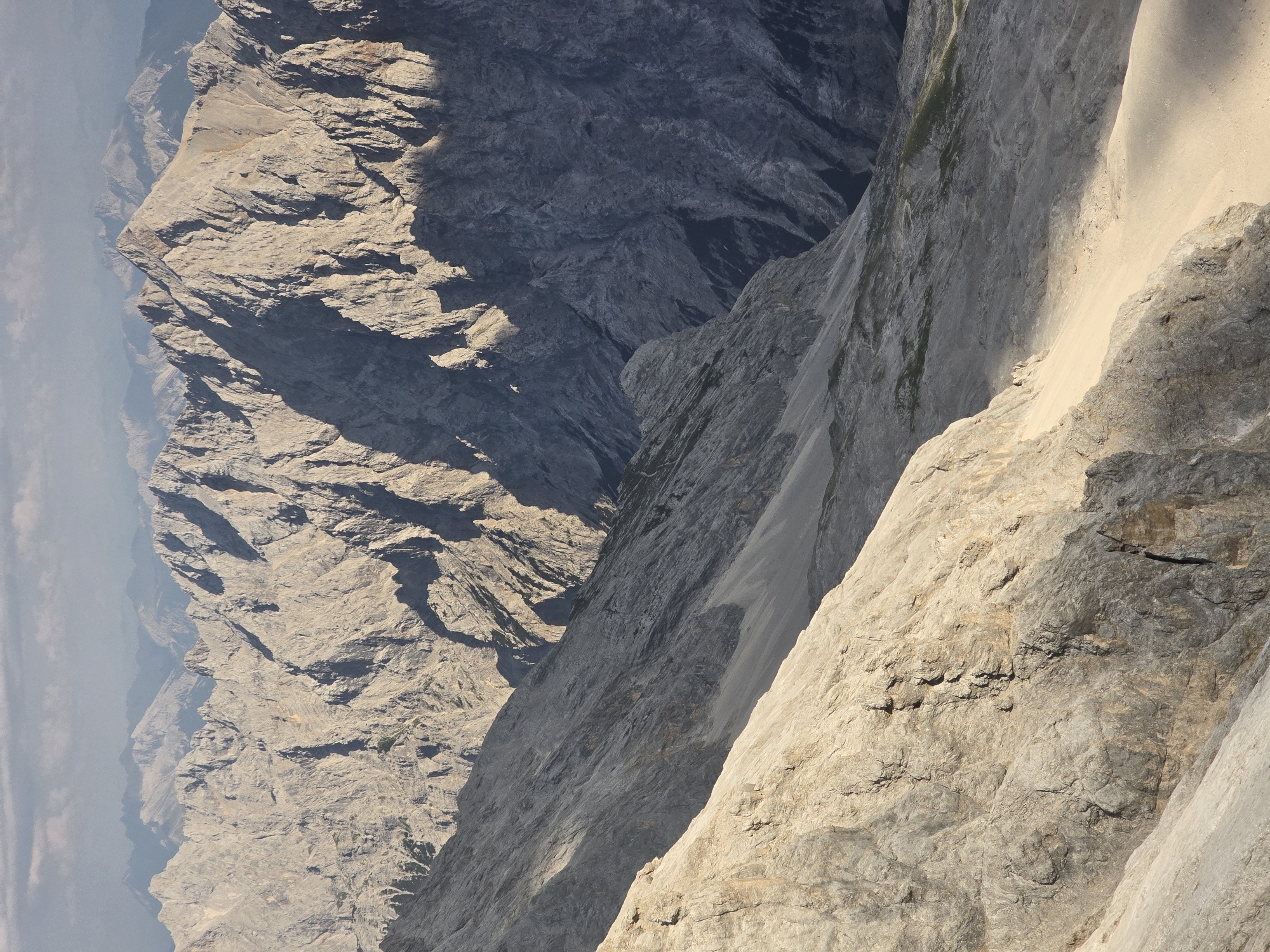 The image depicts a vast, rugged mountain landscape, likely taken from a high vantage point. In the foreground, a steep, sunlit rocky slope dominates the left side of the frame, with a lighter, sandy-colored scree slope descending to the right. Further into the midground and background, the image is filled with layered mountain ranges characterized by sharp, jagged peaks and deep, shadowed valleys. The rock formations are primarily gray and brown, showing evidence of erosion and geological stratification. The sky above is partly cloudy, with a soft, diffused light suggesting either early morning or late afternoon. A small group of figures, appearing as tiny specks, can be observed walking along a discernible path in the midground, indicating human presence in this remote environment. The overall scene conveys a sense of immense scale, natural beauty, and challenging terrain. This landscape is characteristic of the Bavarian Alps and could be within the vicinity of Garmisch-Partenkirchen, Germany, known for its dramatic mountain scenery and hiking trails.