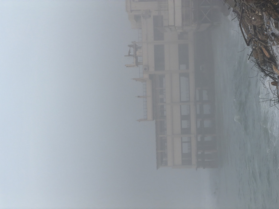 A tall, multi-story building shrouded in dense fog stands adjacent to water. The building's skeletal framework is visible, with rectangular windows and structural supports. Debris, possibly from demolition or natural erosion, is scattered along the water's edge. The overall atmosphere is obscured by the fog, reducing visibility and creating a sense of isolation.