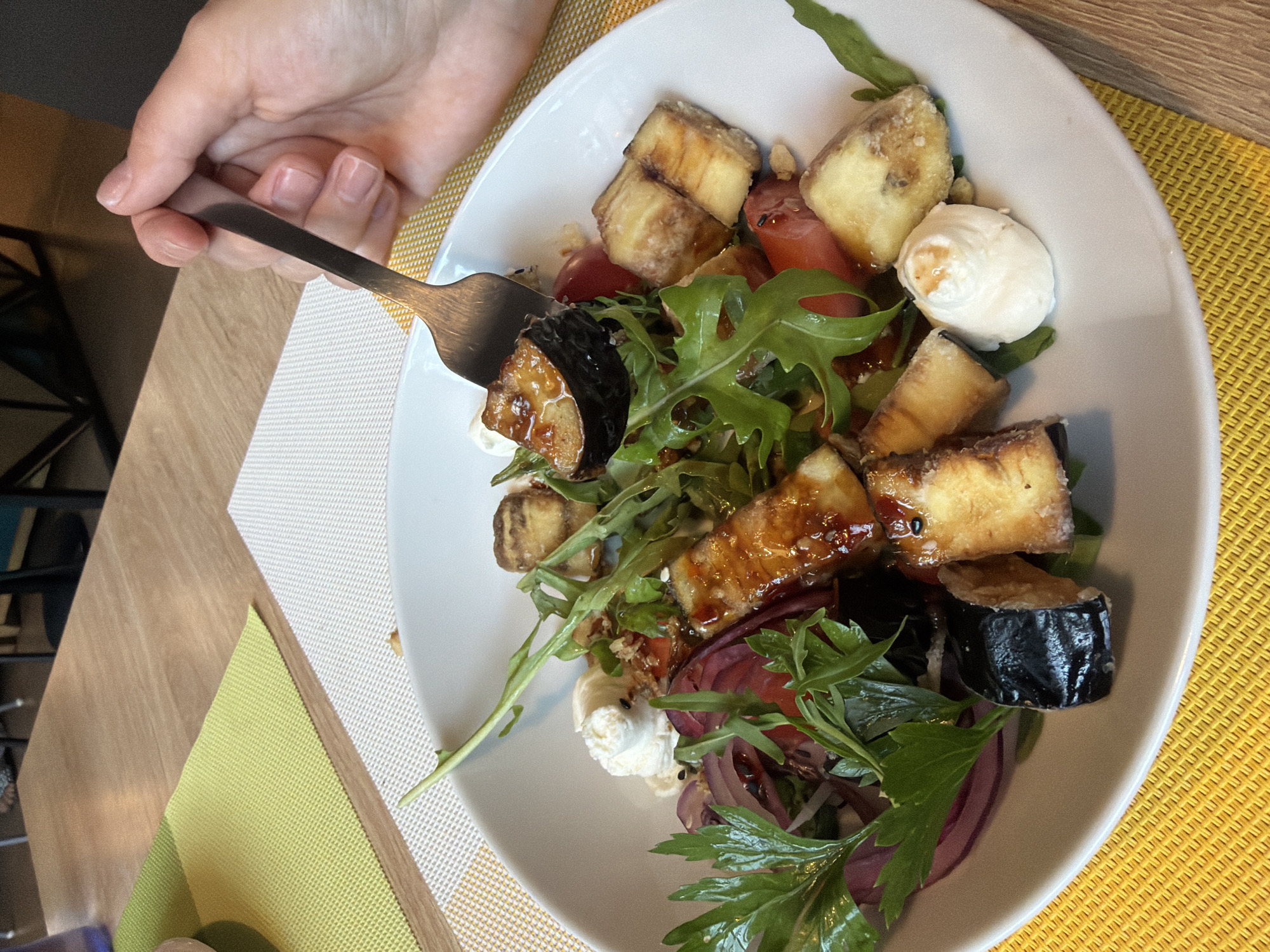 The image shows a close-up of a white bowl filled with a salad. The salad contains pieces of fried eggplant, cherry tomatoes, mozzarella balls, red onion, and arugula. A fork is lifting a piece of eggplant from the bowl. The bowl is on a yellow placemat, which is on a table with a wooden surface. The scene appears to be indoors, possibly in a restaurant. The location is stated to be Chaikovsky, Russia.