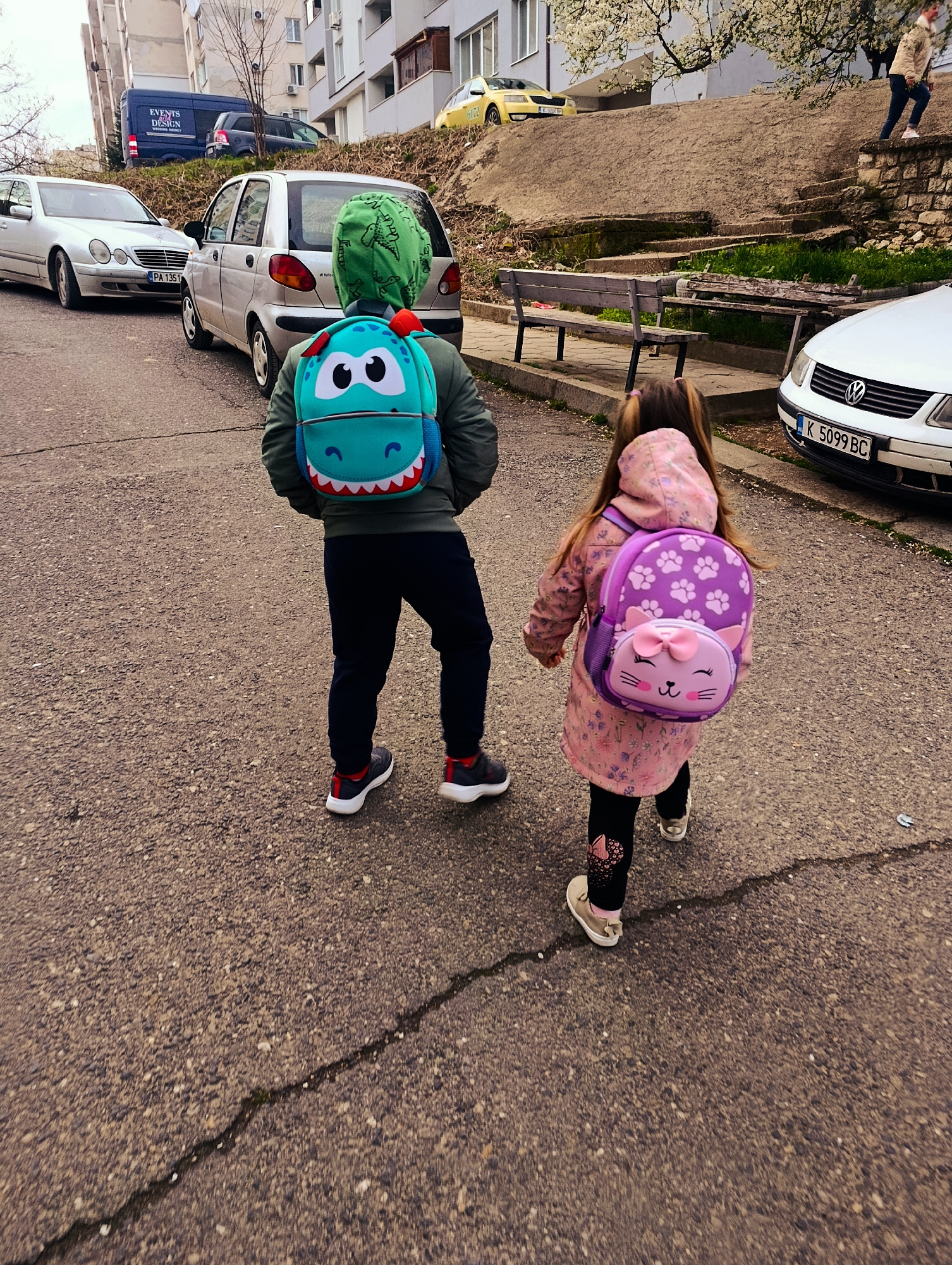 Two young children walk away from the camera on a cracked asphalt street. The child on the left wears a green jacket, dark pants, and dark sneakers with red accents, carrying a blue dinosaur-themed backpack and a green dinosaur-themed hood. The child on the right wears a pink floral-patterned hooded jacket, patterned leggings, and light-colored shoes, carrying a purple cat-themed backpack. A white Mercedes-Benz is parked on the left. A light silver hatchback car is parked beside it. Behind those cars, a dark blue van is parked further uphill. A white Volkswagen Passat is parked on the right side of the street. Benches are along a retaining wall above the right side of the street. Apartment buildings are visible in the background. The license plates read: PA 1351 (Mercedes-Benz), K 5099 BC (Passat), and K 1362 BH (yellow vehicle on hill). The location is likely in the city of Kardzhali, Bulgaria, based on the license plate designations. A person ascends stone steps in the background on the right.