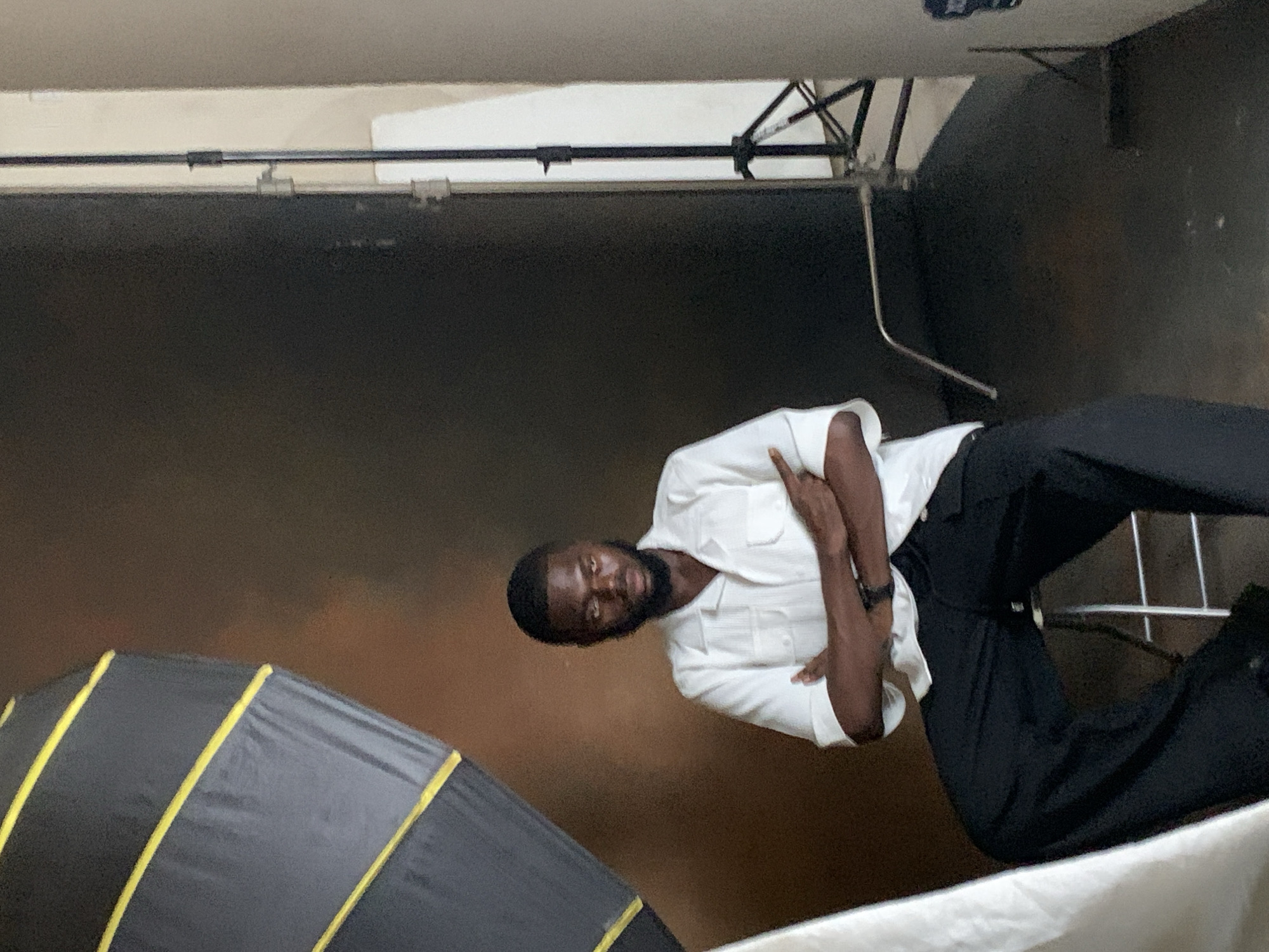 The image depicts a young Black man seated on a stool, facing the camera. He is wearing a white short-sleeved collared shirt with two pockets on the chest and black wide-leg pants. His arms are crossed, and his expression is serious.
The setting appears to be a photography studio. To the left of the man, a large, dark softbox with yellow accents is visible, positioned at an angle. Behind him, the backdrop is a dark, textured brown. To the right, a studio light stand with a white reflector or backdrop is present. The floor is dark.
The image does not contain any information that would allow for its location to be specified in the city of Barikin Jirga Summa, Nigeria.