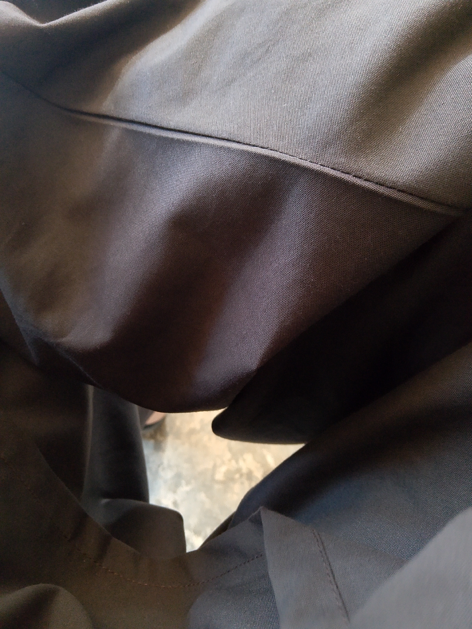 This image shows a close-up view of dark-colored fabric, likely clothing. The fabric appears to be a smooth material, possibly polyester or a blend, with a visible weave. There are folds and creases in the fabric, suggesting it is part of a garment like pants or a skirt. The lighting creates shadows and highlights, emphasizing the texture and form of the material. The background is out of focus and appears to be a light-colored, possibly textured surface, like concrete or pavement. There is no identifiable information or context within the image to suggest a specific location in Mansehra, Pakistan.