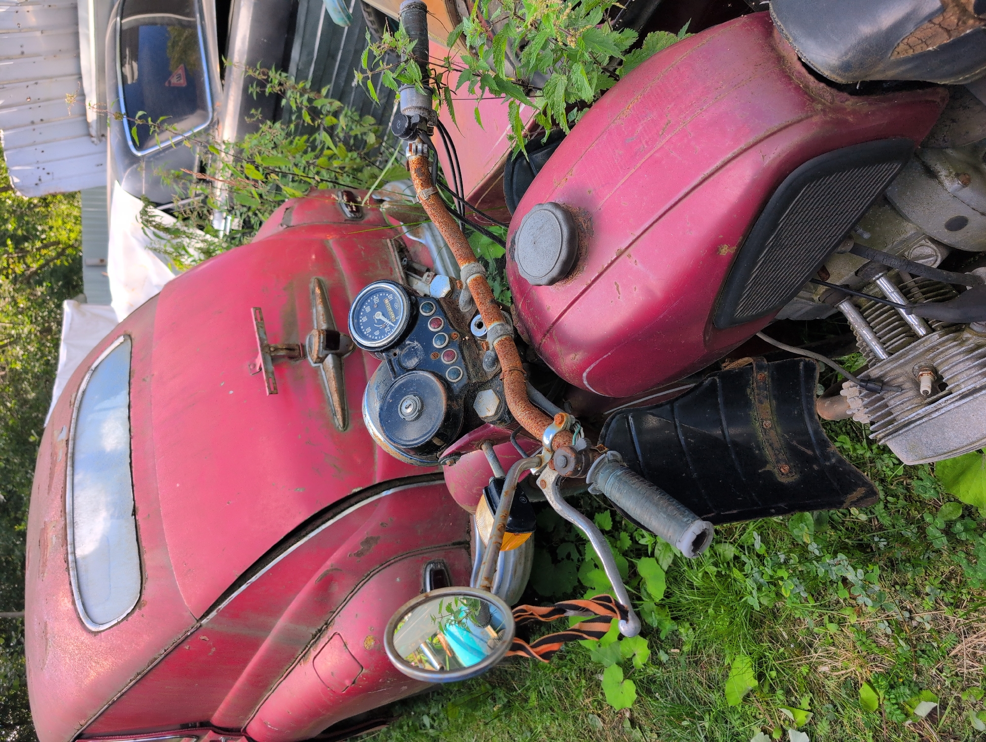 In Novoe Devyatkino, Russia, an abandoned red car and a red motorcycle are visible in an outdoor junkyard or overgrown lot. The car, a vintage model with a rounded rear, is partially obscured by the motorcycle. The motorcycle, also red, has a prominent engine, handlebars with a rusted grip and a mirror attached to the left side. Both vehicles appear weathered and show signs of rust and disuse, with vegetation growing around and on them. The background includes other vehicles and structures, suggesting a storage or disposal area for old machinery. The scene is outdoors, under natural light, with lush green grass and weeds covering the ground.