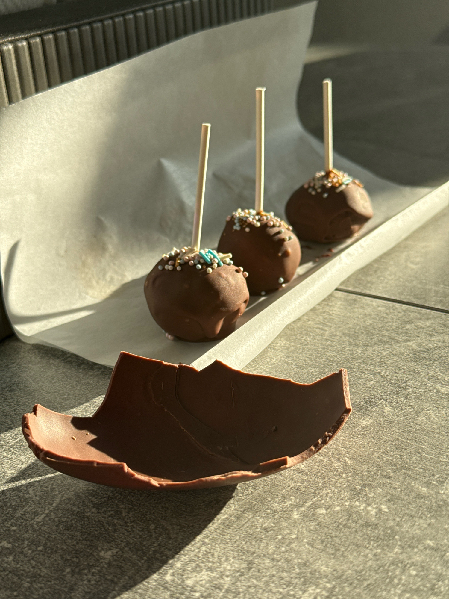 Three chocolate cake pops topped with sprinkles are arranged on a sheet of parchment paper, which is placed on a dark, textured surface. A broken piece of chocolate sits in the foreground. The scene is illuminated by bright sunlight, casting shadows. The image composition suggests an indoor setting with a kitchen appliance visible in the background. The location is not specified in the image, and therefore cannot be determined as Lyubertsy, Russia.