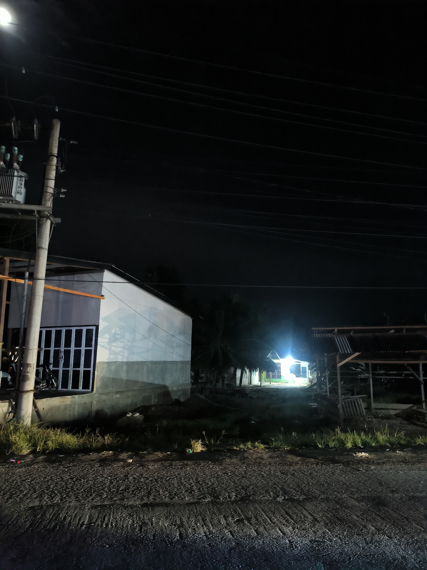 The image presents a nighttime scene in what appears to be a rural or semi-urban area, illuminated by artificial light sources. A rough asphalt or dirt road occupies the foreground, flanked by grass and scattered debris.
On the left side of the frame, a tall utility pole stands prominently, supporting a transformer and multiple power lines that stretch across the dark sky. Below the transformer, part of a white building with a grey foundation and a window featuring white grilles is visible. A dark motorcycle is parked near this building.
Towards the right, an open-sided, unfinished wooden structure with a corrugated metal roof stands, suggesting either a storage shed or an ongoing construction project. Further into the background, a strong, bright light emanates from a distant building, partially obscured by trees, some of which appear to be palm trees. The rest of the scene is enveloped in darkness, emphasizing the artificial light.
The time of day is clearly night, with no natural light visible. The sky is dark and clear. No people are present, lending a quiet, deserted atmosphere to the scene. Indistinct text, possibly East Asian script, is visible on the utility box attached to the pole. The overall impression is one of a calm evening in a developing or residential area.