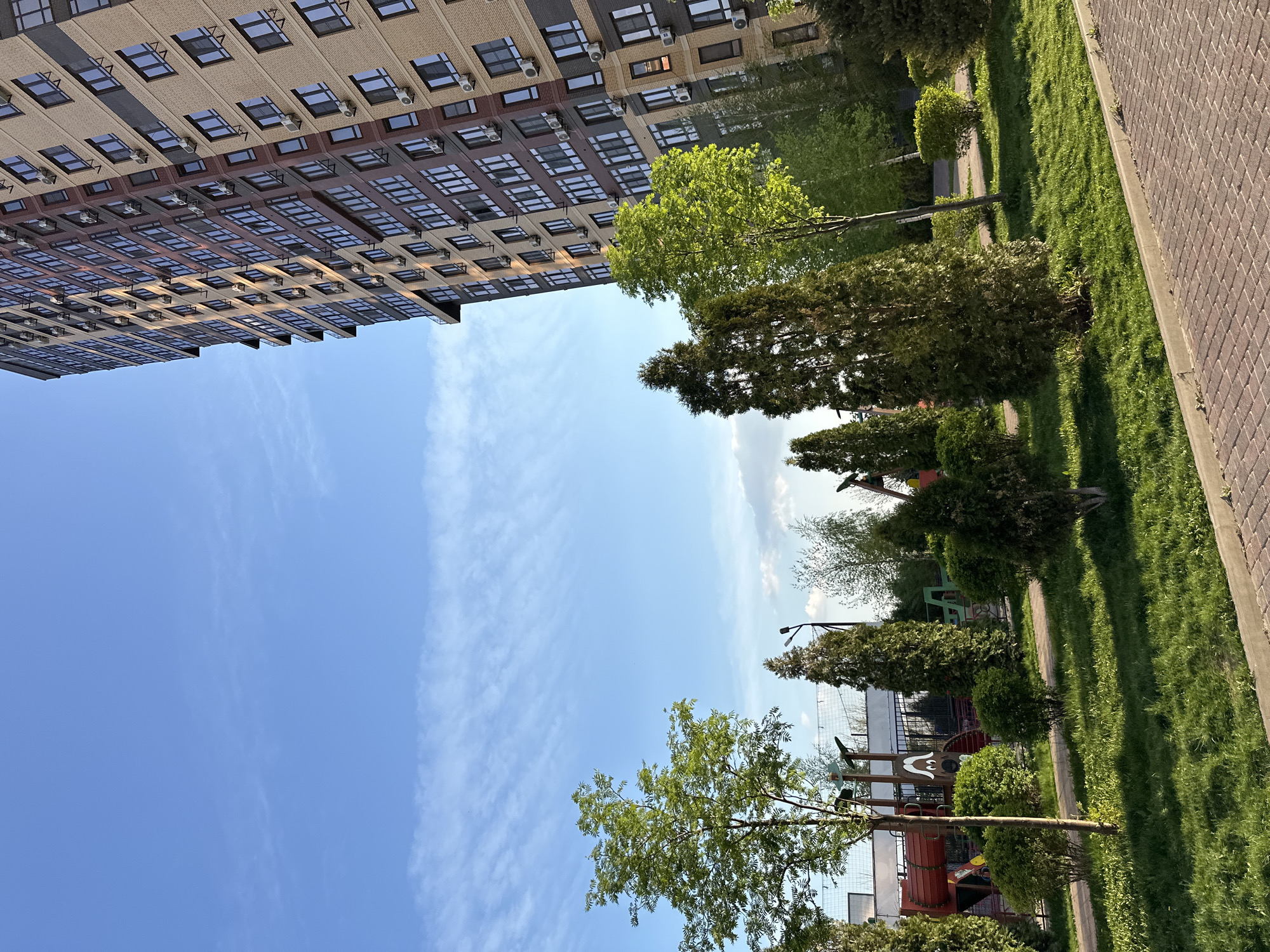 The image presents an upward view of a modern urban scene, likely in a residential area of Rostov-on-Don, Russia. A tall, multi-story building dominates the upper left of the frame. It features a grid-like façade with a mix of brick and gray paneling, and numerous windows, many of which have individual air conditioning units installed. The rest of the image is predominantly blue sky, with some wispy clouds and a streak of thicker cloud cover running vertically. The bottom and right sides of the frame depict a green, well-maintained park area. Various trees and bushes are visible, lining a pathway made of interlocking pavers. In the distance, there is a children's playground with playground equipment visible through the greenery.