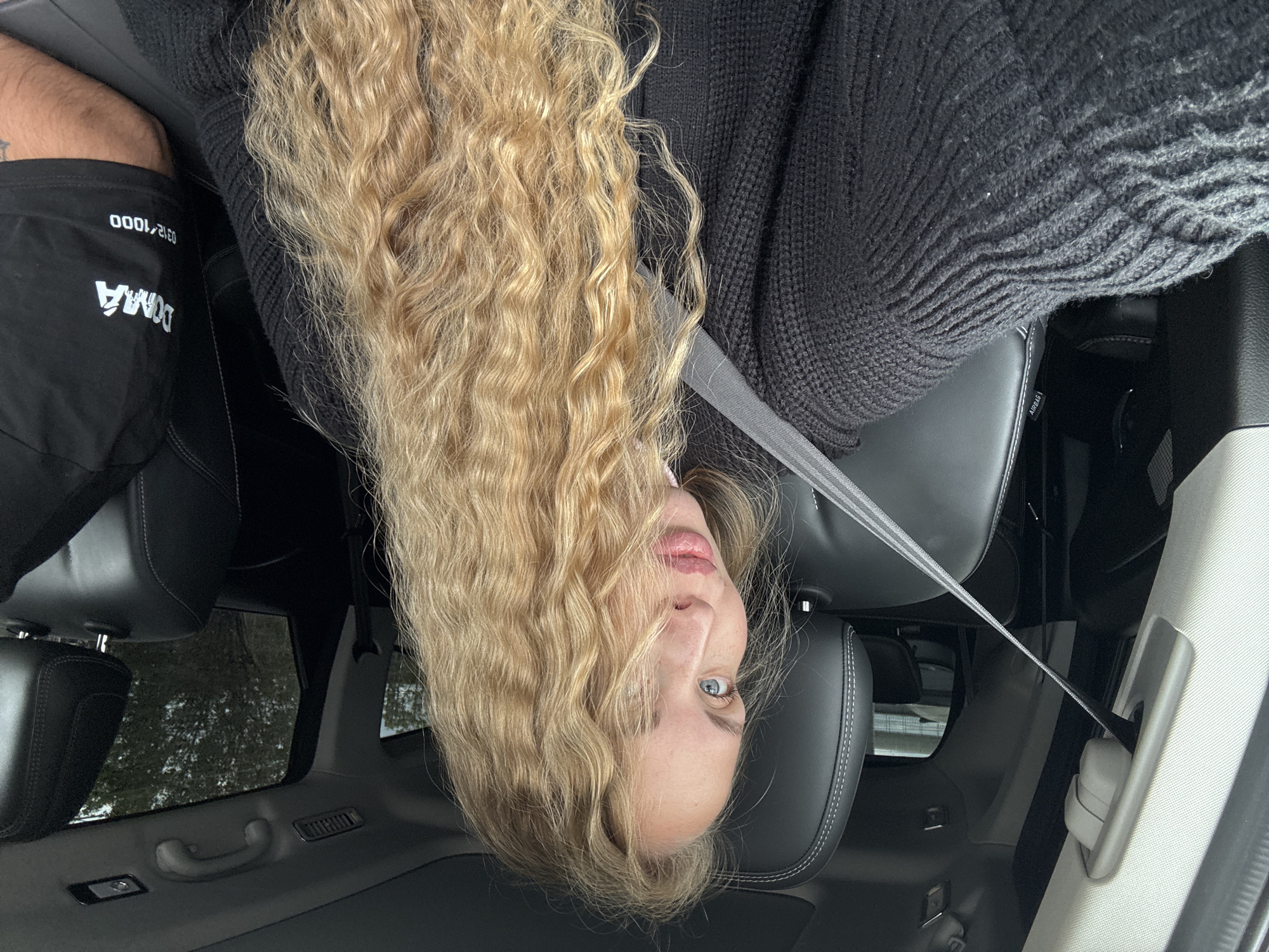 The image shows a person inside a car, presumably the driver, positioned upside down. They are wearing a dark knit sweater and are secured with a seatbelt. The person has long, blonde, curly hair. Part of their face is visible, including light blue eyes and lips. The interior of the car is visible, with black leather seats. Through the car windows, foliage and possibly buildings are visible. A person's upper leg and shorts are visible in the top left of the image. The shorts have "DOMA" printed on them, along with the number "0312/1000". The location in Sochi, Russia, cannot be verified from the image.