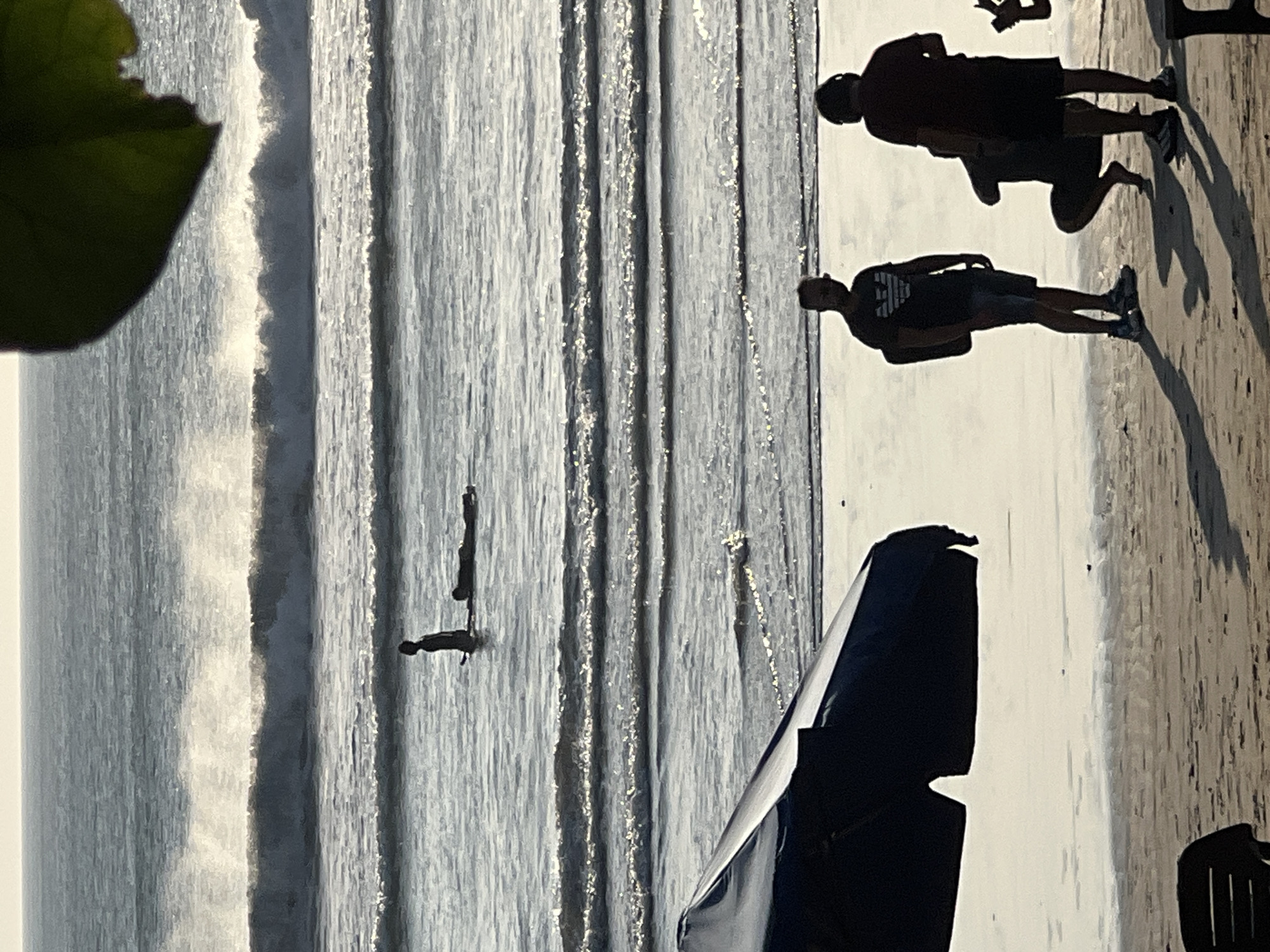 The image shows a beach scene at Kuta, Indonesia. In the foreground, silhouetted figures are visible on the sand. One person stands with a backpack, another is crouching, and a third stands facing away. A large dark umbrella is positioned on the left side of the frame, casting a shadow. In the middle ground, two figures are in the water, one sitting up on a surfboard and another lying prone on a surfboard. The ocean is calm with gentle waves breaking in the background. The sky is bright and hazy, suggesting it is either early morning or late afternoon.
