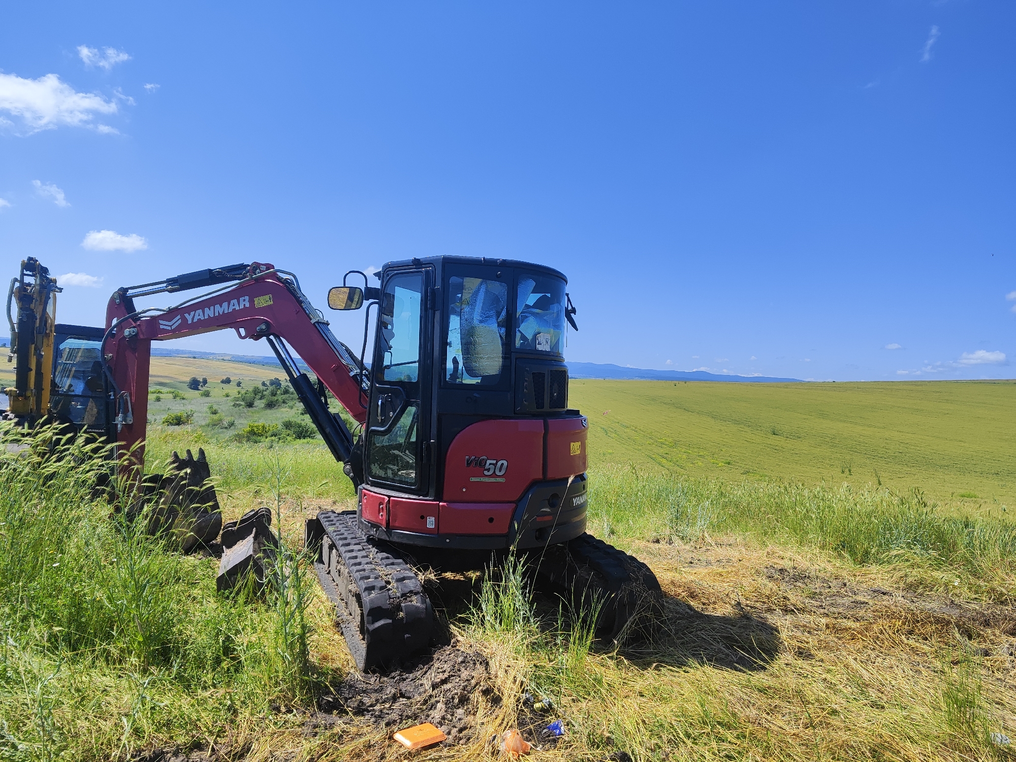 An orange excavator, identified as a Yanmar ViO50, sits idle on a patch of disturbed, grassy terrain under a clear blue sky. The excavator's red and black body is visible from its tracks to its cab, which has tinted windows. To its left, a portion of a yellow excavator is partially obscured by tall green grass. The scene is set against a vast, rolling landscape of green fields, likely agricultural, stretching towards a distant, hazy horizon. Hills are visible in the far distance, and a few scattered trees dot the landscape. The overall impression is one of outdoor work or construction in a rural, agricultural setting.