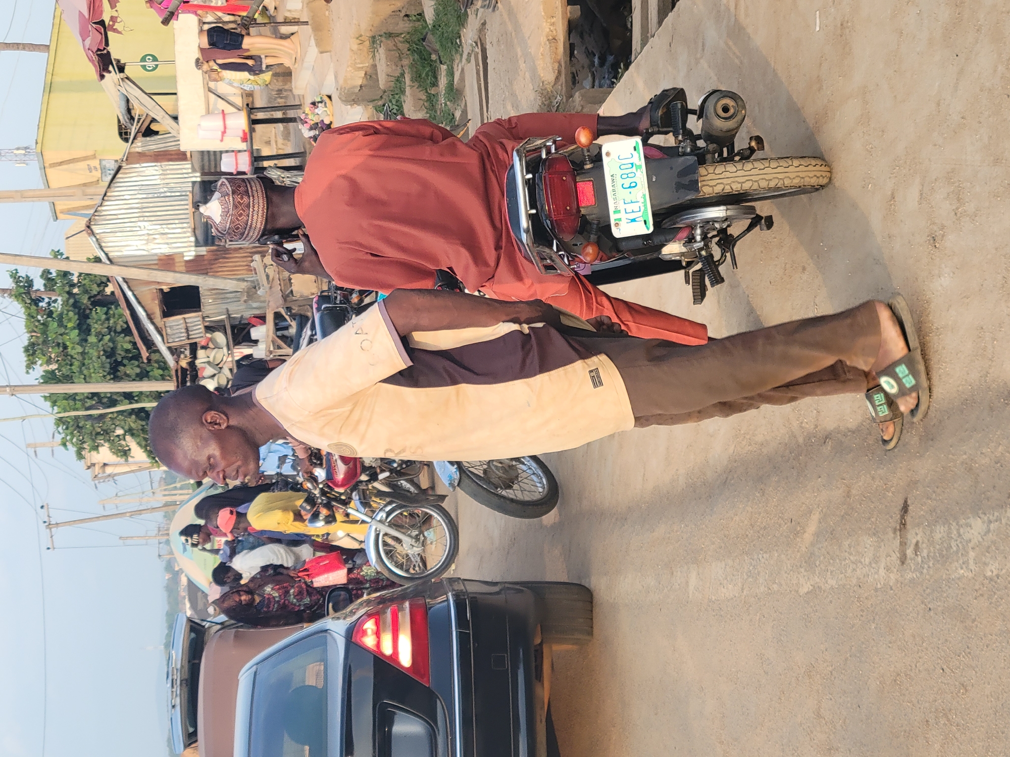 The image captures a vibrant street scene in Keffi, Nigeria, with people and vehicles engaged in daily activities. The setting appears to be a bustling area, possibly a market or residential street, under bright daylight.
In the foreground, a man stands on the right side of the frame, facing left. He wears a light beige and dark brown two-toned short-sleeved top, brown trousers, and green patterned slides. His head is shaven. To his left, another man is seated on a motorcycle, oriented slightly towards the left. This man is dressed in a reddish-brown long-sleeved tunic and a distinct brown and white patterned traditional cap. His back is largely towards the viewer.
The motorcycle he is riding features a prominent green Nigerian license plate that reads "NASARAWA" at the top and "KEF 680C" below it, indicating its registration in Nasarawa State, which aligns with the given location context of Keffi. The motorcycle itself is dark, with chrome elements and a red taillight.
Further into the background, a crowd of people is visible, some gathered near what appear to be makeshift stalls or roadside vendors. White plastic containers are stacked on a table in this area. Other motorcycles and cars, including a black sedan partially visible in the bottom left, line the street. The buildings in the background are modest, featuring corrugated iron roofs and simple construction, characteristic of local commercial or residential structures. Green foliage from trees is also present in the distant background. The street surface is paved.
The scene depicts a typical day with individuals moving around, standing, and waiting, suggesting a casual, everyday atmosphere without a specific organized event. The lighting indicates it is daytime, possibly morning or late afternoon.