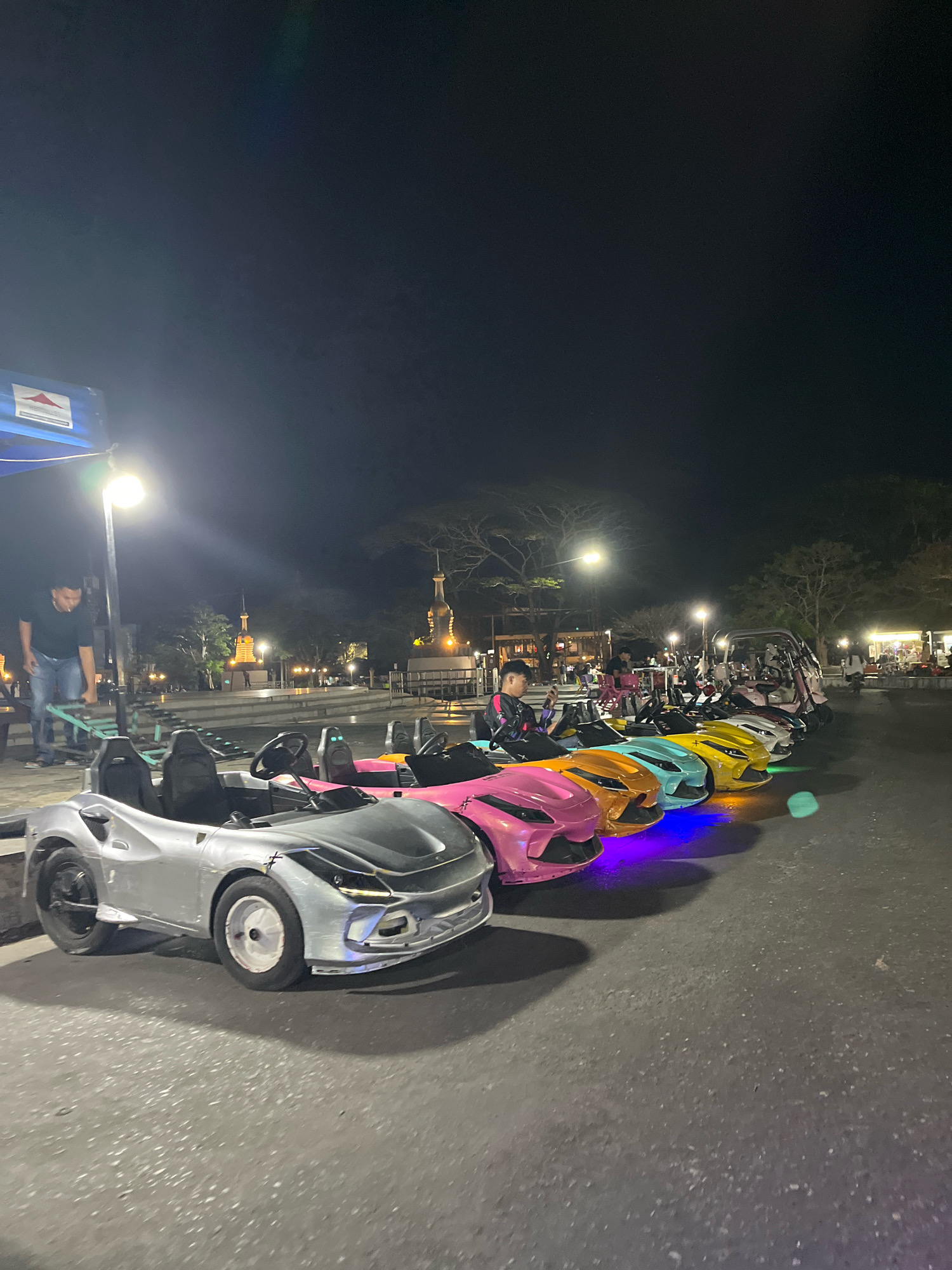 In Kendari, Indonesia, at night, a line of colorful, small electric cars, resembling sports cars, are parked on an asphalt surface. The cars are in various bright colors including silver, pink, orange, teal, and yellow, and some are illuminated with colored underglow lights. In the foreground, a silver car with black seats and a black steering wheel is prominently featured. A person in a black shirt and blue jeans is visible to the left, standing near a blue canopy and a lamppost, appearing to be interacting with some equipment. In the background, several other people are seated in some of the cars or at tables, and there are illuminated buildings, trees, and streetlights, suggesting an outdoor entertainment or recreational area. The sky is dark and clear.