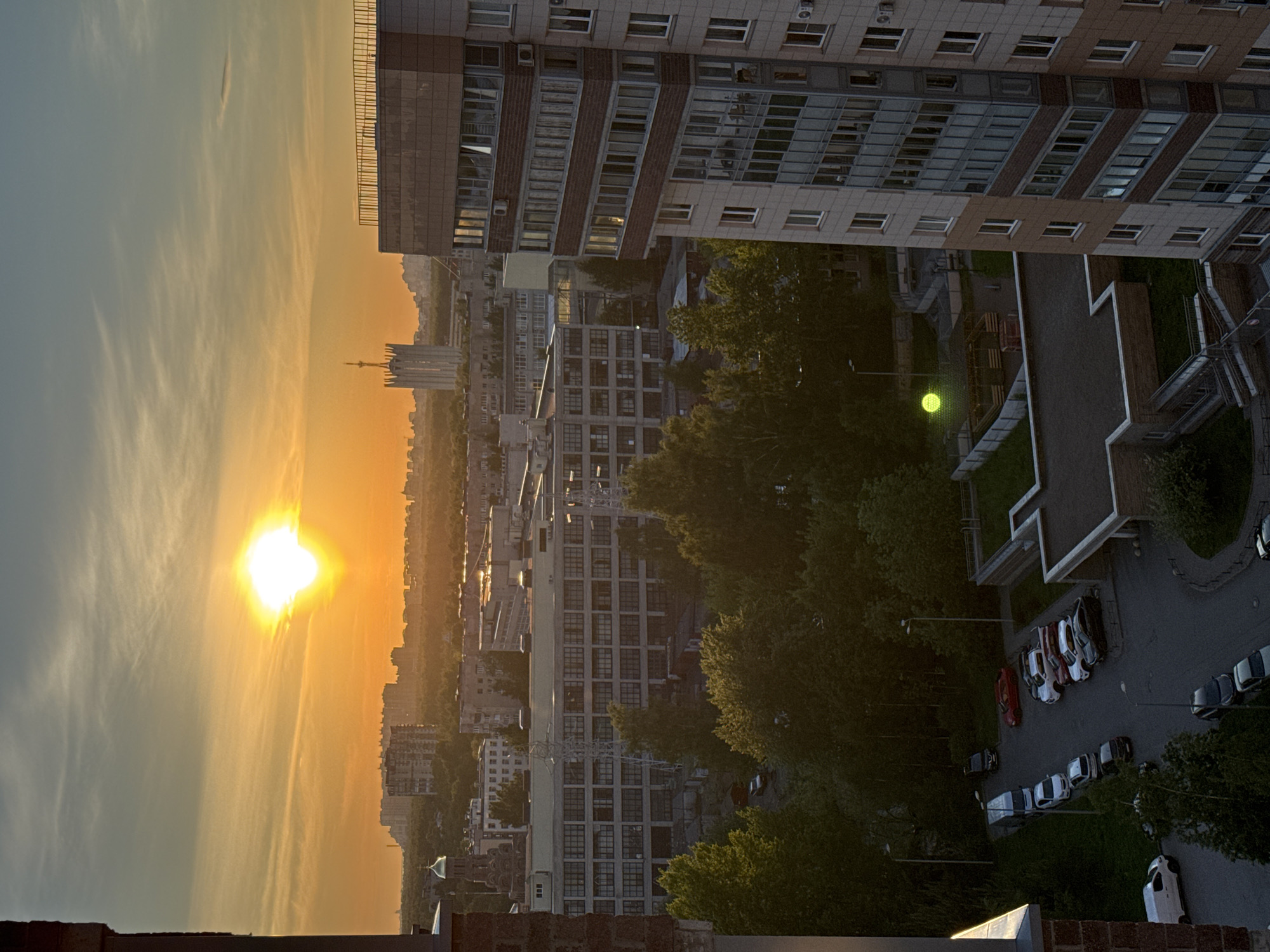 This image depicts a cityscape at sunset, likely Ladozhskoe Ozero, Russia, based on architectural styles and landscape features. In the foreground, a tall residential building dominates the right side of the frame. A courtyard below features trees, a playground, and parked cars. Adjacent to the building, other multi-story structures recede into the distance. On the left, the setting sun casts a bright orange glow across the sky and buildings, creating silhouettes of distant structures. Wisps of clouds are visible in the sky. A brick structure occupies the bottom center of the frame.