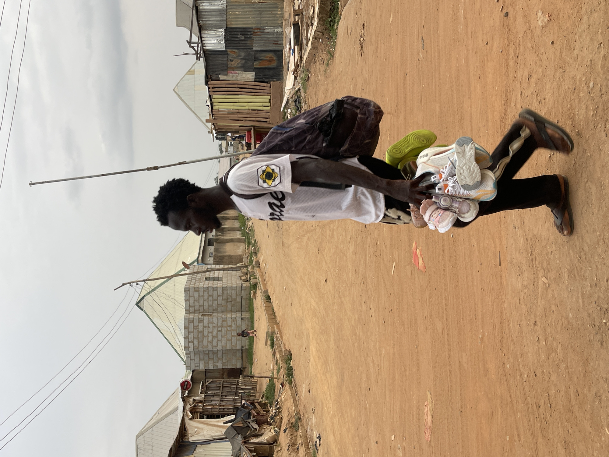A man is walking along a dirt road in Karmajiji, Nigeria. He is carrying a bag on his back and several pairs of shoes in his hand. He is wearing a white t-shirt with black leggings and sandals. The environment includes dwellings constructed of mismatched and incomplete materials, the sky is grey, and the ground is dusty and mostly bare. Another individual is slightly visible, walking in the opposite direction in the distance.