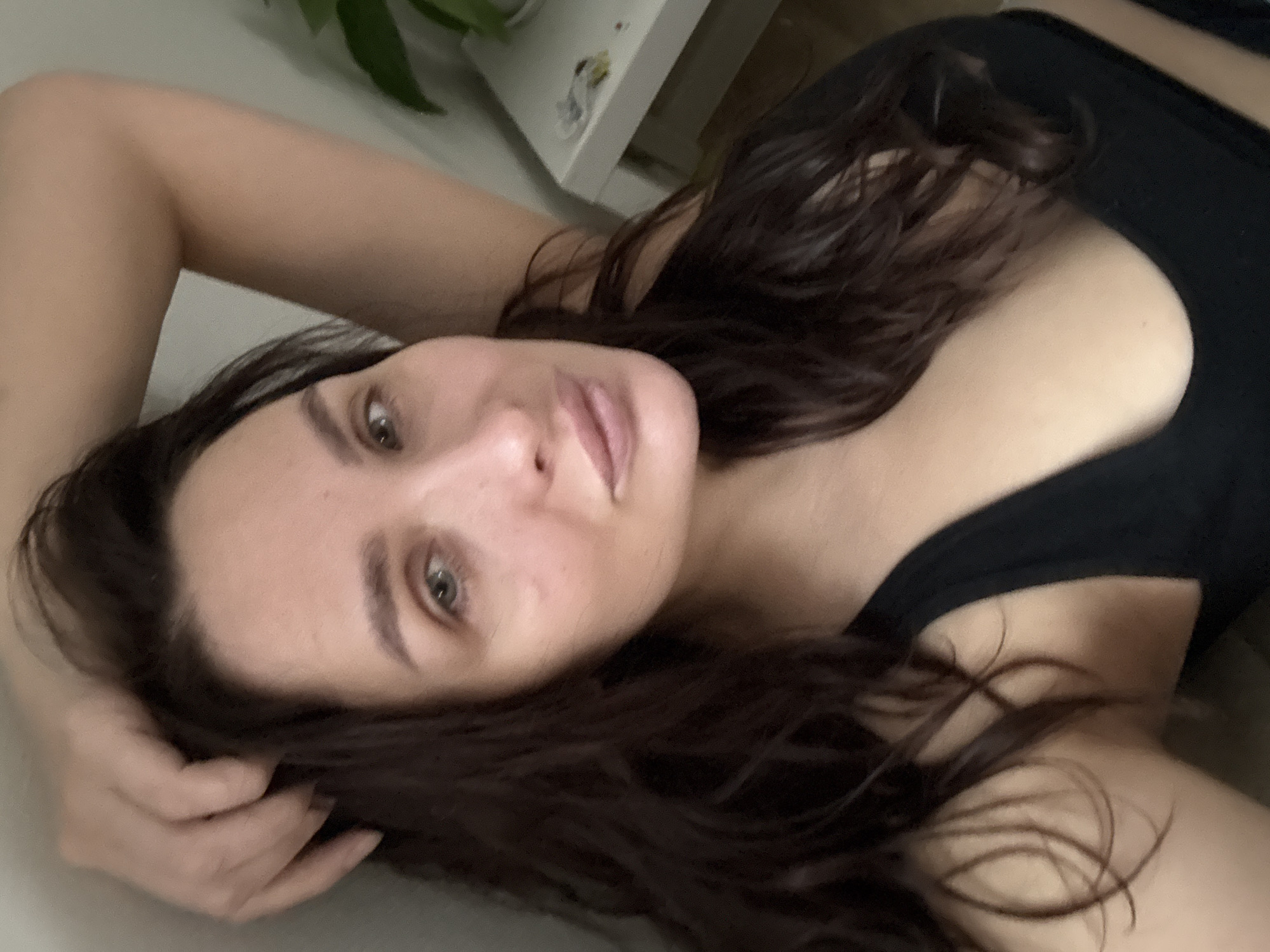 An indoor, close-up, high-angle photograph taken in Chaikovsky, Russia, depicts an adult female. She has fair skin, dark brown wavy hair, and greenish-hazel eyes. Her lips are full and exhibit a light pinkish-nude color. The subject is wearing a black top or undergarment. Her left hand is raised, with fingers lightly touching her hair above her forehead. Her right arm is bent and positioned behind her head, largely out of view. She gazes directly at the camera with a neutral expression. The background consists of a plain, light-colored wall. To the right, a segment of a white rectangular table or dresser is visible, on which indistinct small items, including a light-colored crumpled object, a dark object, and a reddish mark, are present. A portion of a green leafy plant is situated in the far-right background.