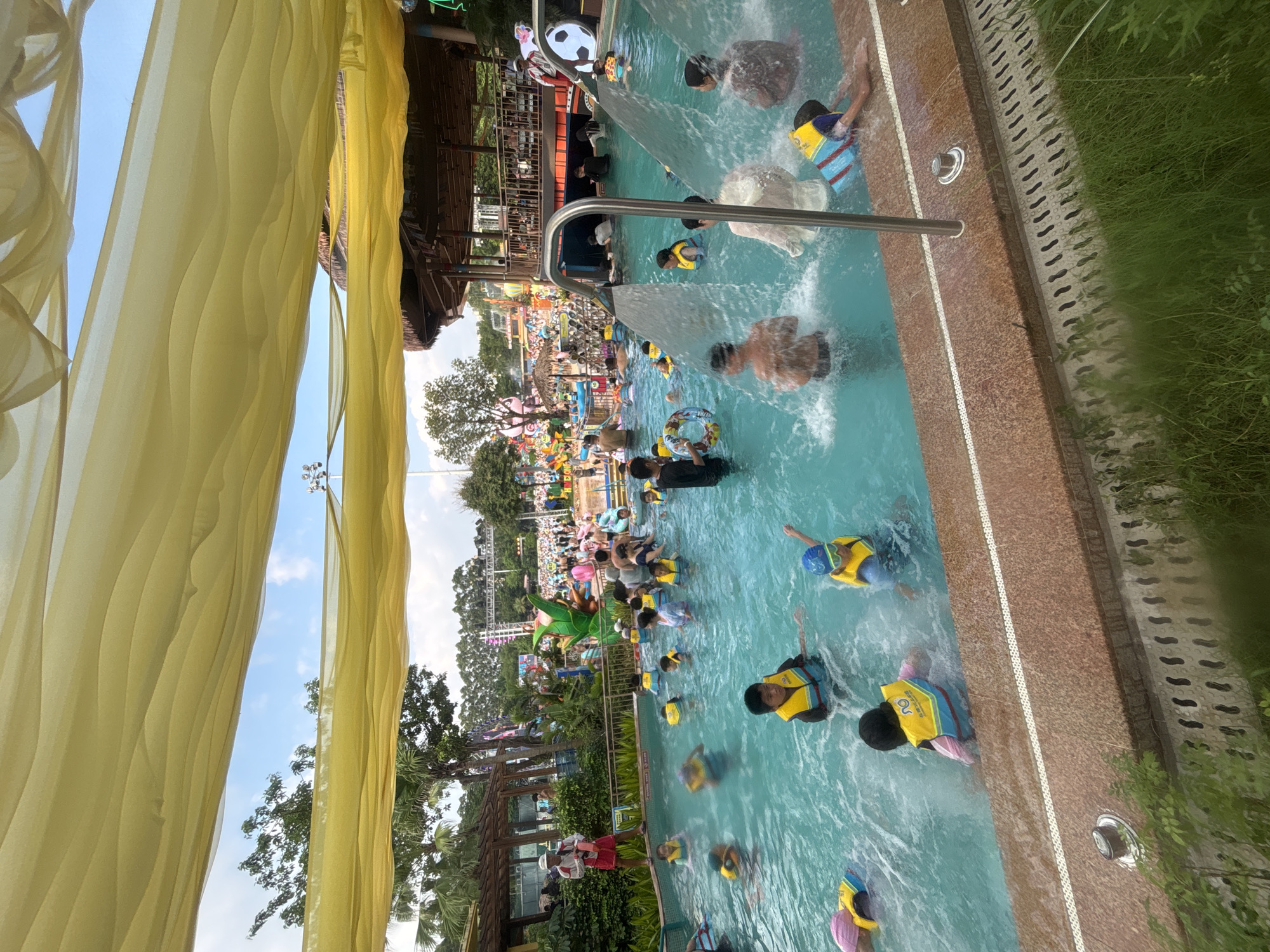 In Panyu District, China, a vibrant water park is bustling with activity under a large, draped yellow canopy. Numerous individuals, many wearing yellow life vests, are enjoying the swimming pool. Children are visible playing in the water, some with inflatable rings, while adults are also present, supervising and participating. Water cascades from overhead structures into the pool, creating a refreshing atmosphere. In the background, a multi-level building with a thatched roof and a lively scene of park attractions and trees suggest a festive and recreational environment. The scene is captured from a low angle, looking up towards the canopy and the sky beyond.