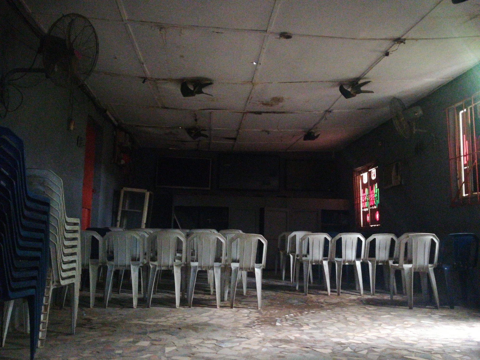 The interior of a dimly lit room in Lagos, Nigeria. A collection of white plastic chairs are arranged in rows. Stacked blue plastic chairs are on the left. There are windows with red bars and decorative stained glass. Ceiling fans are mounted on the ceiling. The ceiling appears damaged. The floor is stone tiled.