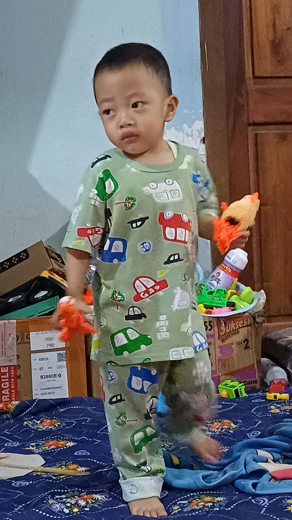 A young child, appearing to be of Southeast Asian descent, stands in what appears to be a room in Wonokerso, Indonesia. The child is wearing a light green pajama set adorned with a repeating pattern of cartoon cars and trees. The child's left hand is holding an orange stuffed toy, and the child's right hand is holding a yellow and orange plush toy. In front of the child's right side, a small collection of toys, including a green toy vehicle and a purple bottle with a spray nozzle, is visible. The background consists of a light blue wall with a shadow cast upon it, and a wooden cabinet. The child is standing on a blue blanket with a patterned design. Several cardboard boxes are present behind the child, with some displaying shipping labels.