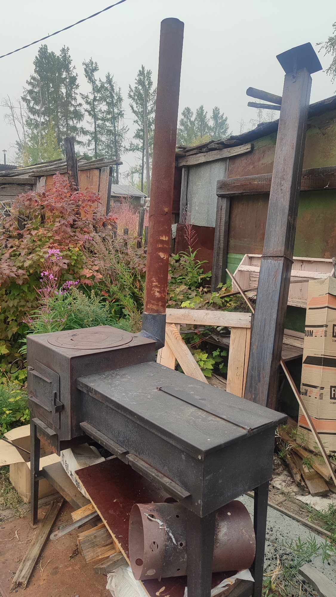 This is a medium shot, eye-level photograph of an outdoor setting in Mirny, Russia. The scene is dominated by a large, black metal stove with a rectangular cooktop and a smaller square section on the left, likely the firebox. A thick, rusty metal chimney extends vertically from behind the stove. To the right of the stove, a tall wooden post with a smaller wooden structure attached to its top is visible. Behind the stove and chimney, there are various structures made of wood and corrugated metal, some appearing weathered and dilapidated. Lush green foliage and bushes with red and pink flowers are growing around the stove and the structures. Several cardboard boxes, some with orange and white branding, are stacked near the wooden post. The ground is covered in dirt, debris, and scattered wooden planks. The sky is overcast and hazy. The overall impression is one of a rustic, possibly abandoned, outdoor area with a functional stove at its center.