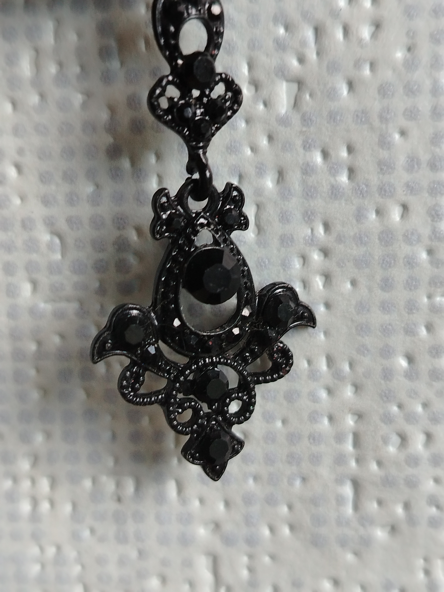A black ornamental pendant with dark stones is centered in the frame against a textured white background. The pendant features intricate metalwork with leaf-like and circular embellishments. The background appears to be a close-up of a fabric or textured surface with a repeating pattern of raised dots. The lighting is uniform, highlighting the detail of the pendant. There are no people or other objects visible in the image. The scene is indoors with a shallow depth of field, focusing on the pendant.
