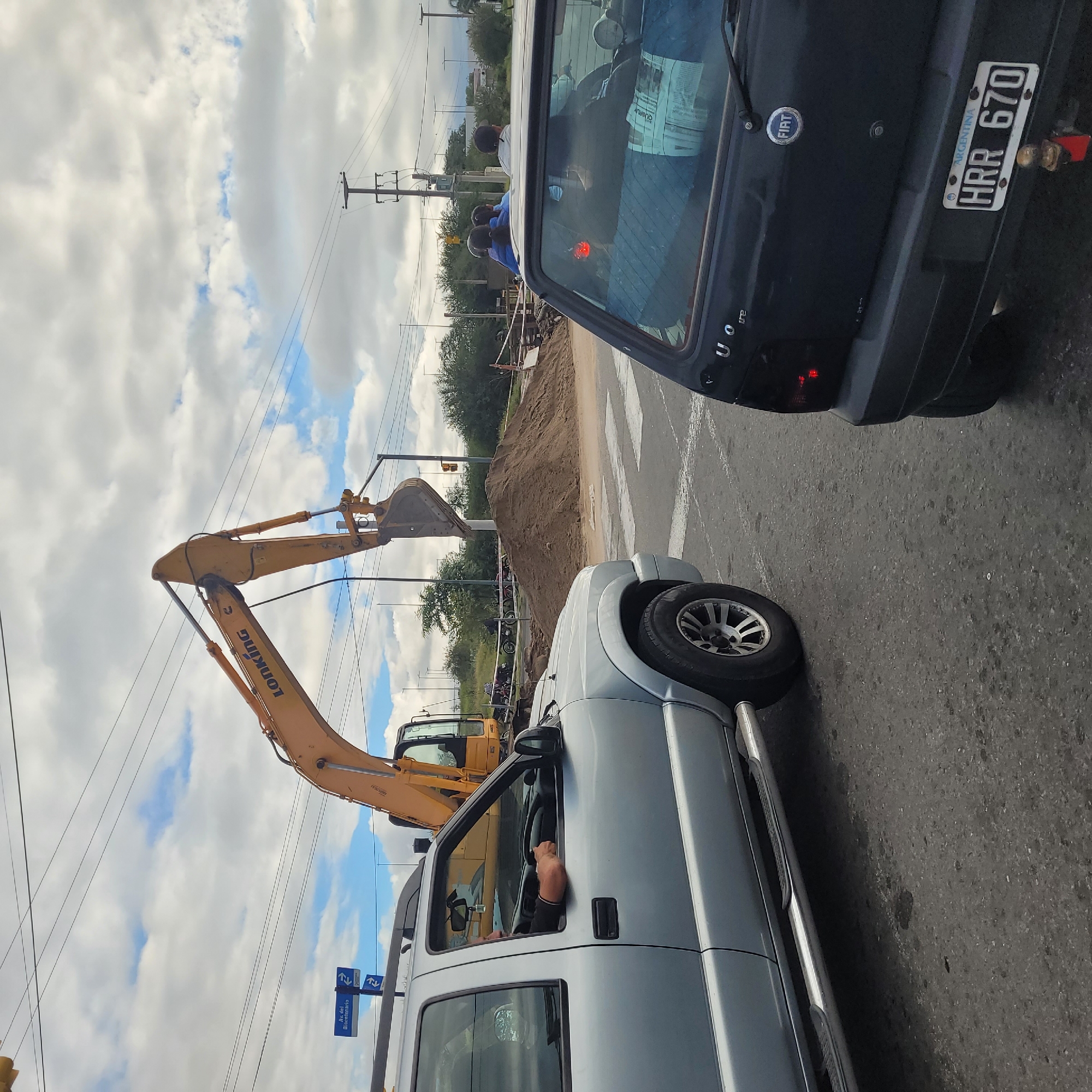 The image depicts a construction scene on a street in what appears to be San Fernando del Valle de Catamarca, Argentina, based on the license plate visible on a black Fiat Uno. A yellow LONKING excavator is positioned near a pile of dirt on a pedestrian crosswalk. A silver pickup truck is to the right of the frame with an unidentified driver inside. A group of individuals wearing blue attire and hard hats are observed in the background behind the pile of dirt. Power lines and street signs including an Avenida del Bicentenario sign are visible in the scene.