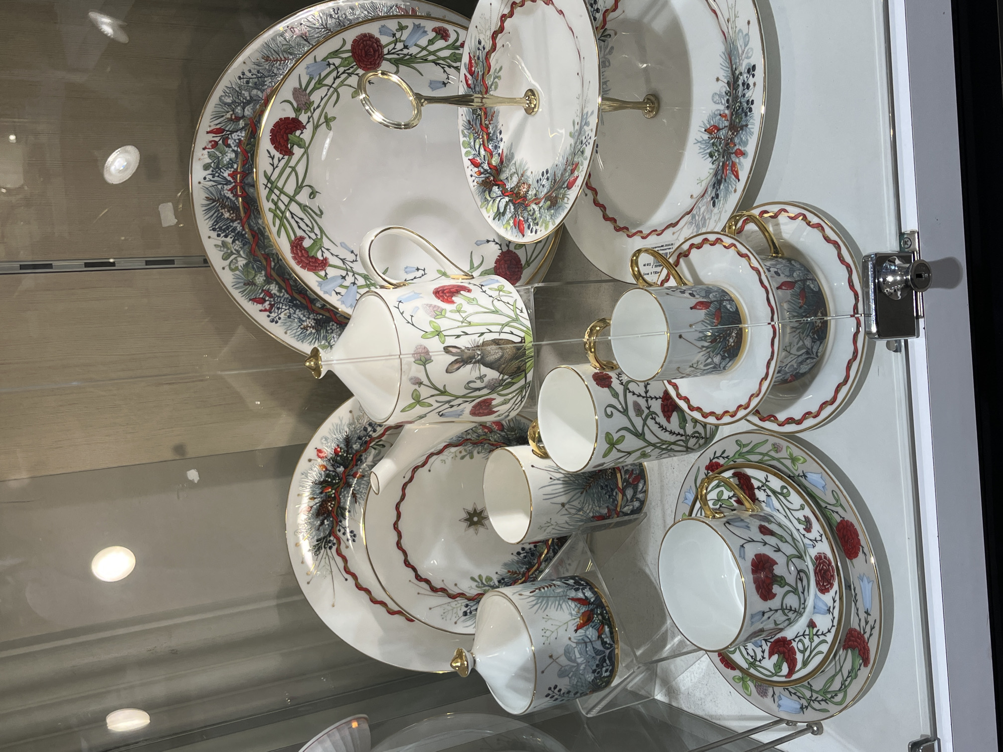 A collection of fine china is displayed behind glass. The china includes plates, cups, and a two-tiered serving dish. The pieces are white with intricate floral and foliage designs in red, blue, and green, accented with gold trim. One cup features a rabbit design. The display appears to be inside a store or museum, possibly in Saint Petersburg, Russia, given the style of the china and the presence of glass display cases. The lighting is bright, reflecting off the glass and polished surfaces of the china. The scene suggests a display of valuable and decorative tableware.