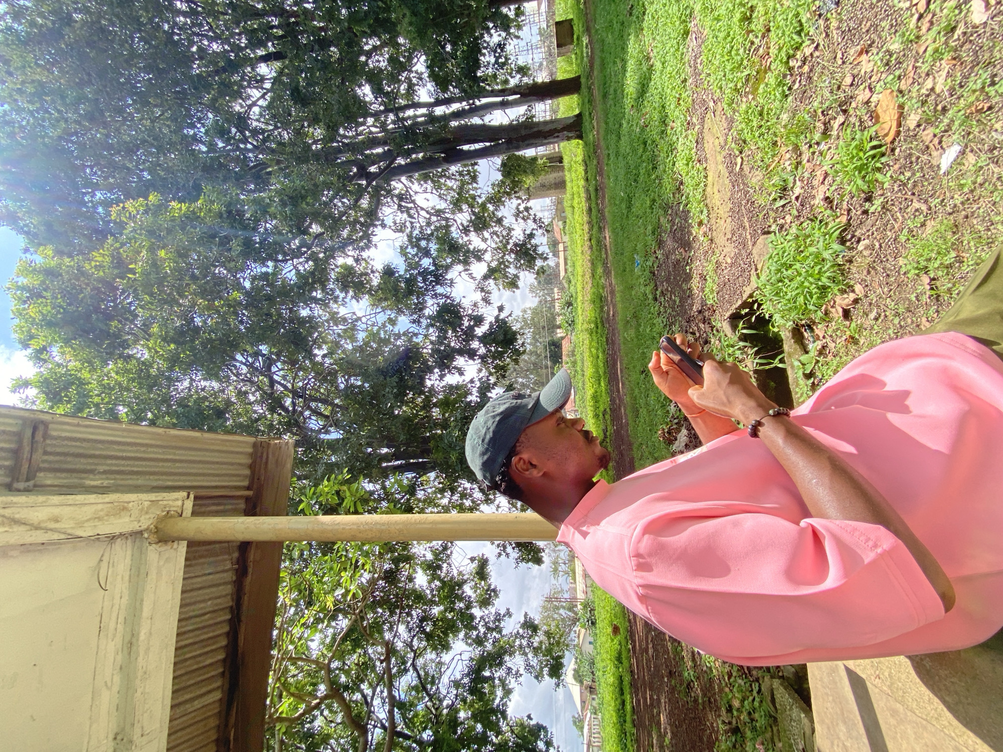 In Bukuru, Nigeria, a young Black man sits outside under a corrugated metal awning, looking down at his mobile phone. He is wearing a bright pink t-shirt and a dark green baseball cap. The man's left arm is adorned with a beaded bracelet. The area appears to be a park or natural setting, with a lush green lawn, trees, and a dirt path. In the background, a few buildings and structures are visible, suggesting the presence of a nearby settlement or industrial area. The sky is bright blue with scattered clouds, and sunlight streams through the trees, creating a dappled lighting effect.