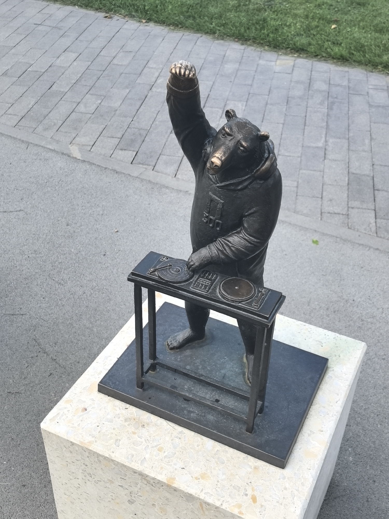 A bronze statue of a bear in a tracksuit is shown in a city park in Perm, Russia. The bear stands on a square granite pedestal with its right arm raised and its left hand on a DJ turntable. The bear is wearing a hooded jacket with an emblem on the chest. The statue is located on a paved walkway next to a grassy area.
