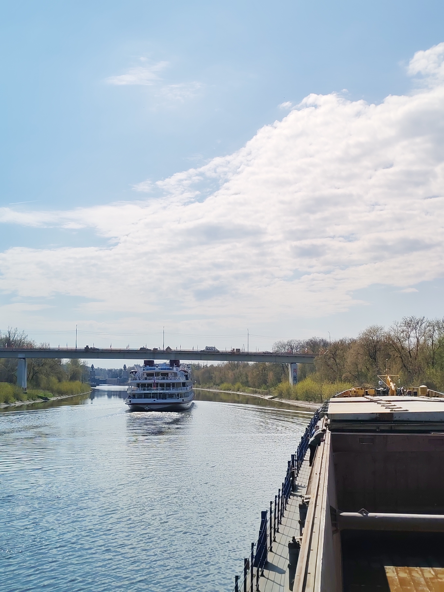 The image shows a waterway in Yakhroma, Russia. A passenger cruise ship is sailing towards the camera, passing under a bridge carrying vehicle traffic. On the right side of the frame, a barge is also navigating the waterway. The sky is partially cloudy. Trees line the banks of the waterway. In the distance, beyond the bridge, architectural landmarks are visible.