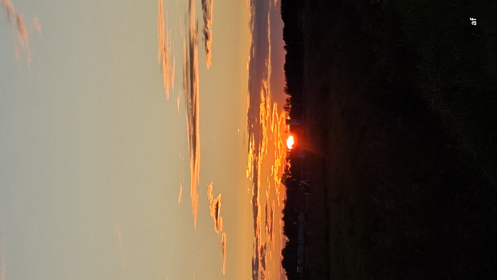 The image captures a sunset over a rural landscape, possibly on the outskirts of Tiskre, Estonia. The sky is painted with warm hues of orange and yellow, transitioning to a soft blue at the top. Wispy clouds are illuminated by the setting sun, which appears as a bright, glowing orb just above the horizon. Silhouetted against the vibrant sky is a dark, treeline that suggests a forested area or a line of trees bordering fields. In the foreground, a dark, indistinct field or grassy area is visible, also silhouetted against the twilight sky. A few faint, dark shapes that could be structures or figures are barely discernible in the distance, but are not clear enough to be identified. The overall impression is one of natural beauty and tranquility at the end of the day.