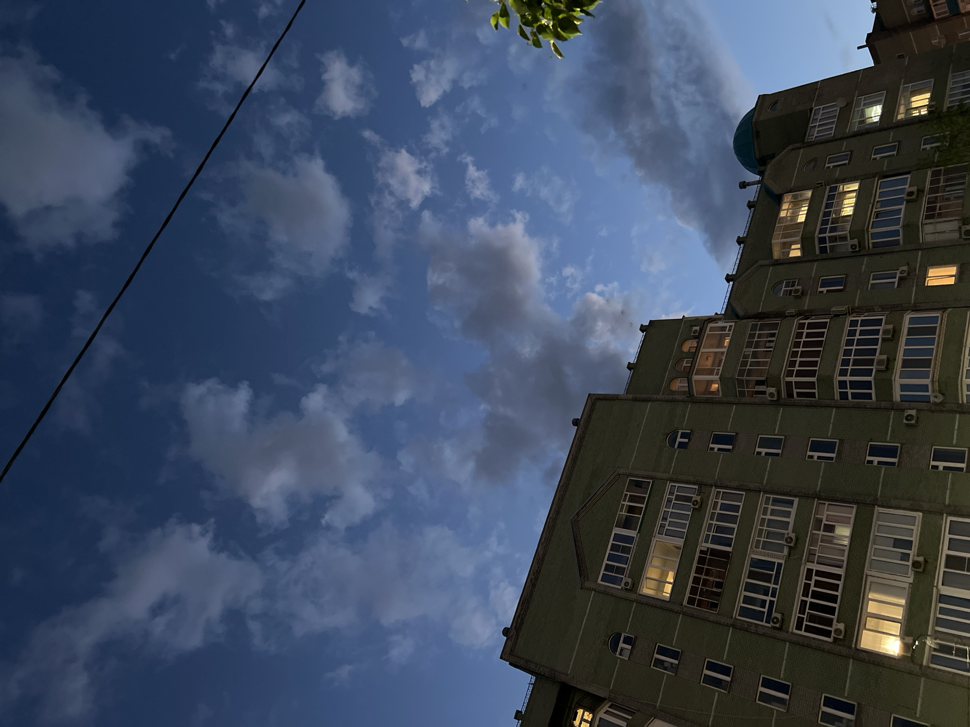 The image shows a residential building in Kazan, Russia, likely at dusk or dawn. The building is tall and appears to be made of green brick. Many windows are visible, some with lights on inside, suggesting that people are home. The sky is a deep blue with scattered clouds, some of which are dark and appear heavy. A thin black wire runs diagonally across the top portion of the image. In the lower right corner, a small section of green leaves from a tree is visible, adding a touch of nature to the urban scene. The perspective of the photo is upward, looking towards the sky and the upper floors of the building.