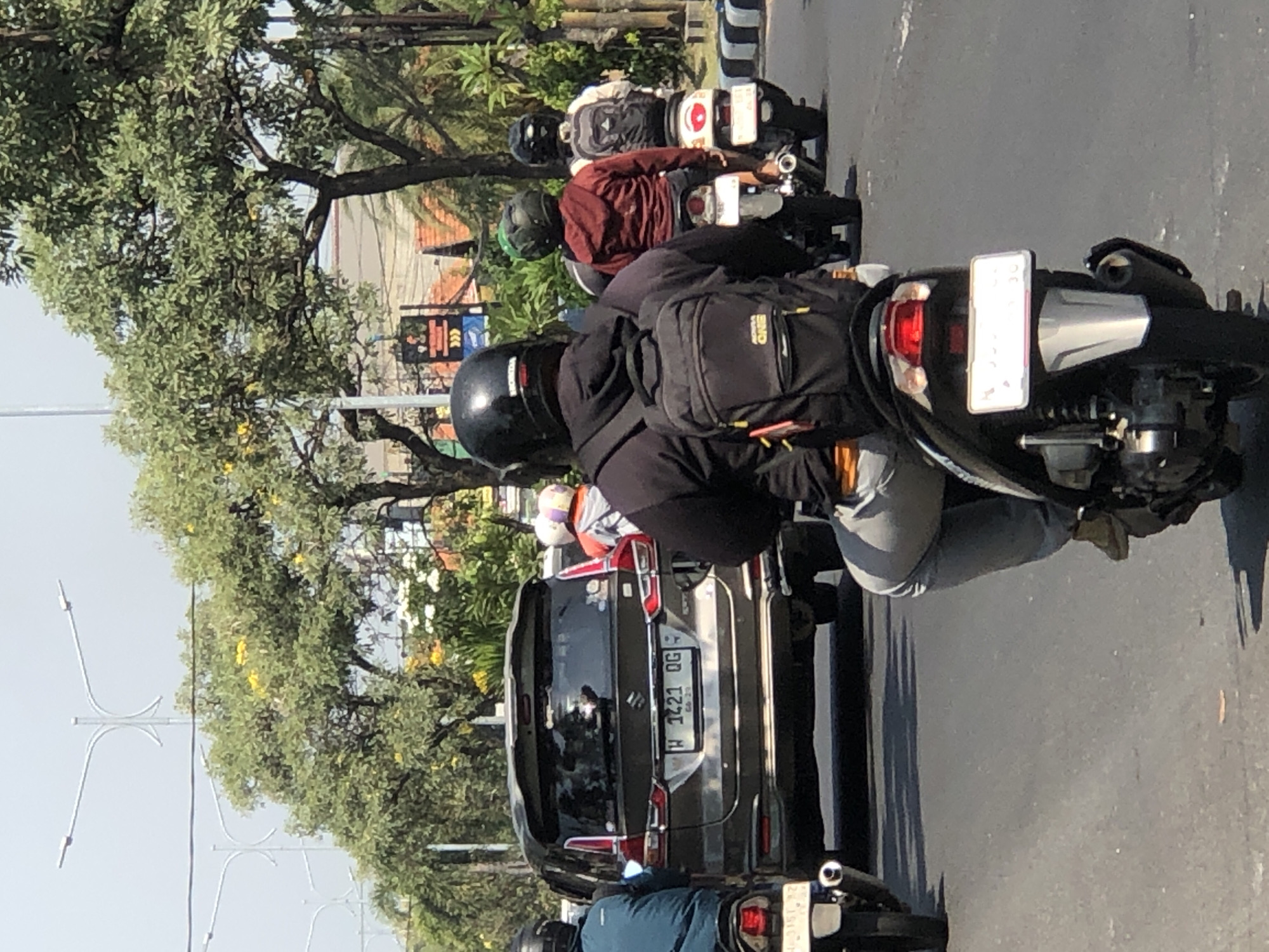 The image shows a street scene in Larangan, Indonesia, with several motorcycles and a car. In the foreground, a person wearing a black helmet and a black hoodie with a backpack is riding a motorcycle. Behind them, another motorcycle with a rider wearing a red hoodie and a black helmet is visible. Further back, a dark-colored SUV is driving in the same direction. The road is paved and lined with trees and some buildings in the background. The sky is bright and appears to be daytime. There are streetlights visible in the distance. The overall impression is of a typical urban traffic scene.