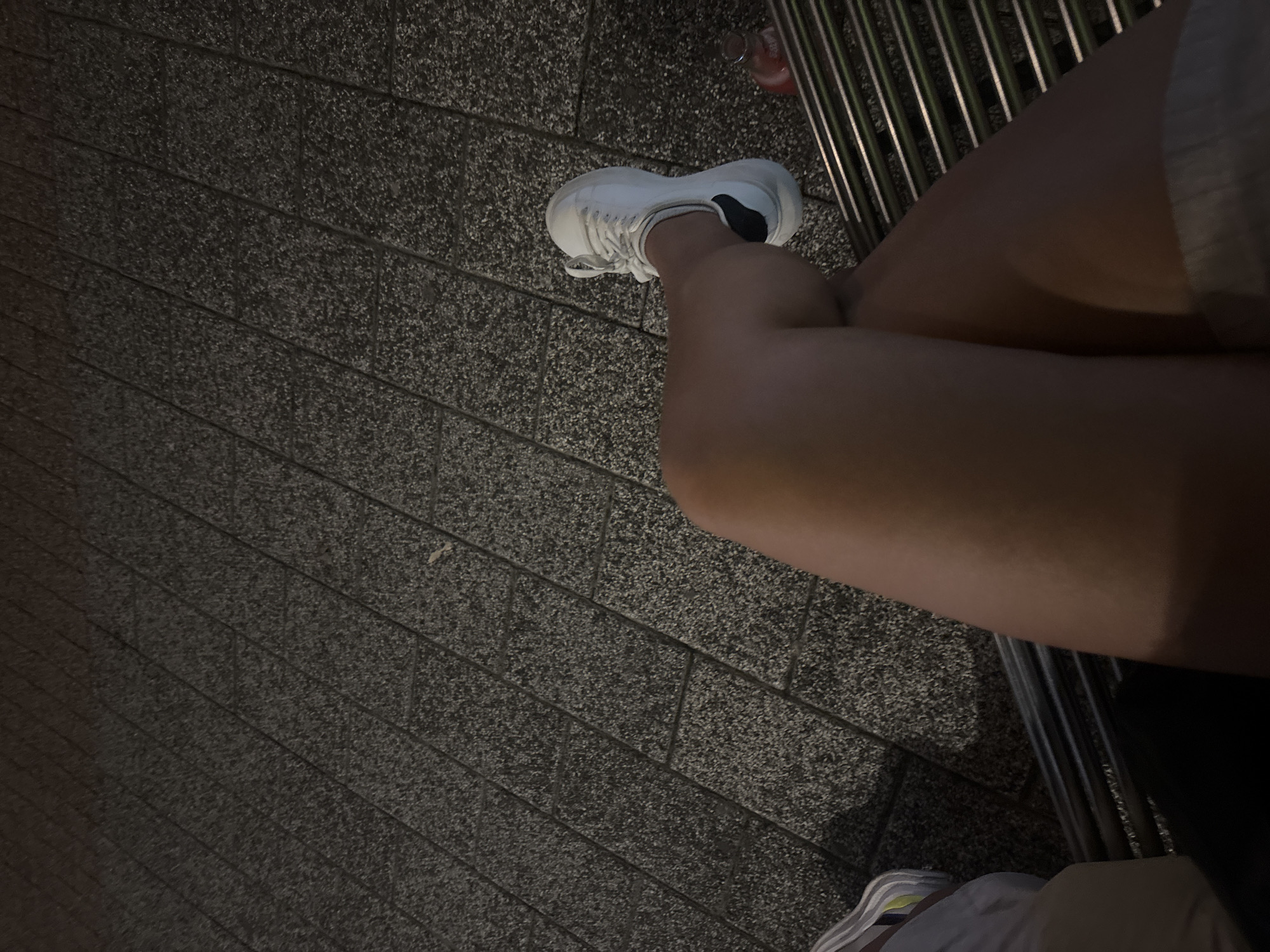 The image shows a close-up of a person's legs and feet, sitting on a bench outdoors at night. The ground is paved with dark, speckled tiles arranged diagonally. The person is wearing white sneakers with black accents. Their legs are crossed, with one knee visible above the other. A portion of a metal bench with horizontal slats is visible in the lower right. A small, red bottle with a silver cap is also present on the bench. The overall lighting suggests it is nighttime. The location is the city of Plovdiv, Bulgaria, though specific landmarks are not identifiable from the provided view.