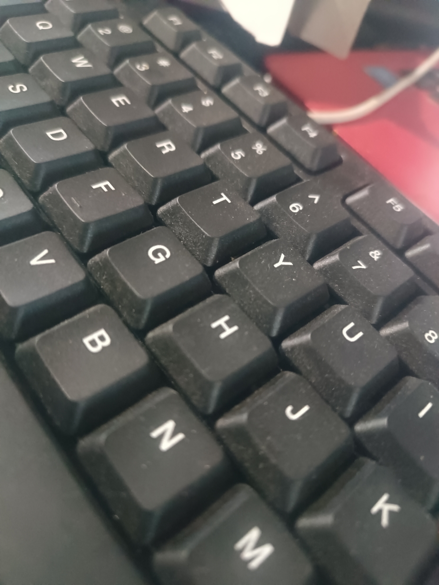The image is a close-up, high-angle shot of a black QWERTY keyboard. The keys are worn, with visible dust. The letters on the keys are white. Some of the keys visible are Q, W, E, R, T, Y, U, I, A, S, D, F, G, H, J, K, Z, X, C, V, B, N, M. The background is blurred and appears to be a red surface with a white cable.
There is no information in the image to suggest this scene is located in Tabanan, Indonesia.