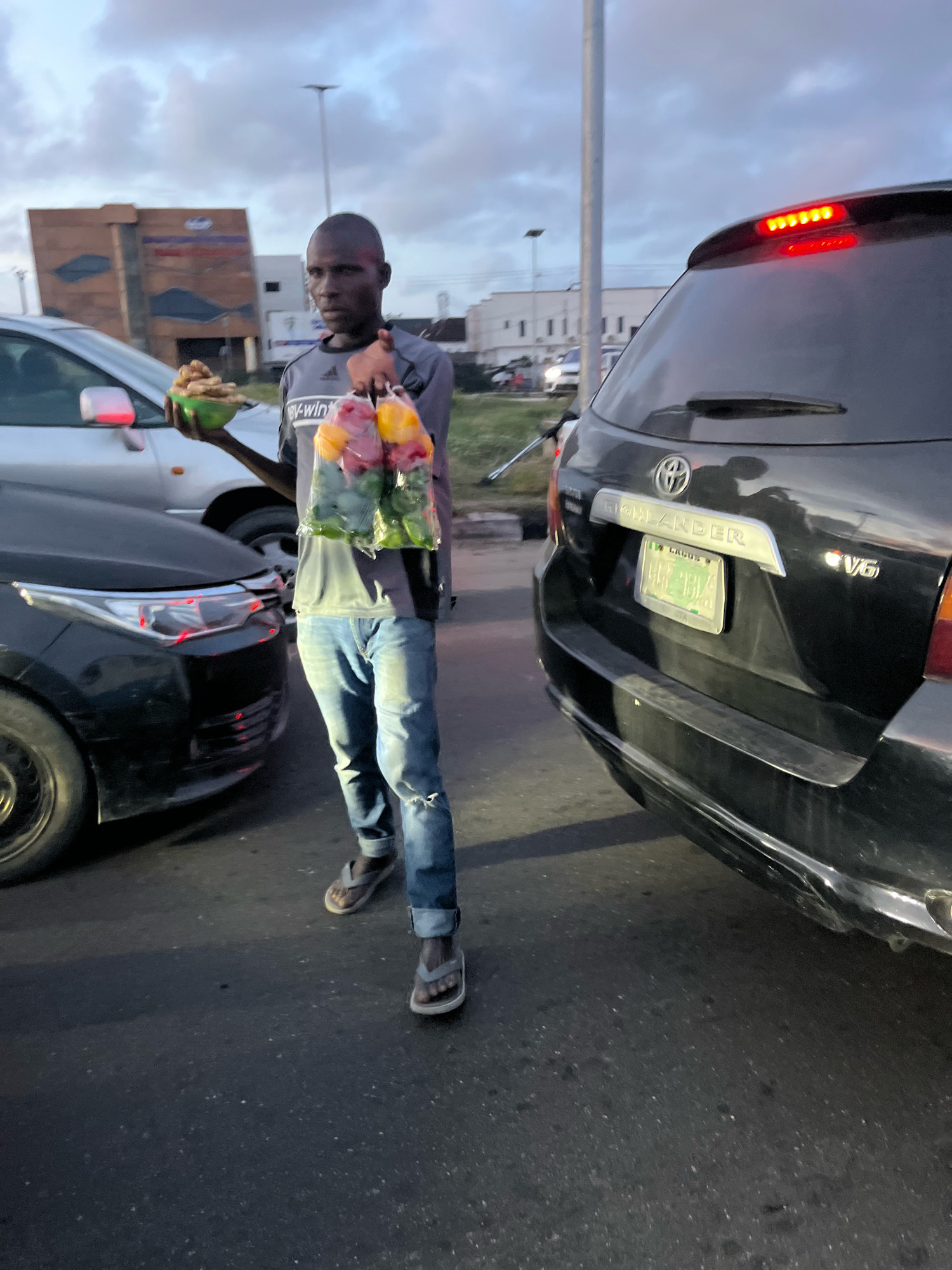 A man stands on an asphalt road in what appears to be the city of Lekki Peninsula II, Nigeria. He is holding a green bowl in his left hand and a plastic bag filled with bell peppers in his right. The man is wearing a grey t-shirt and blue jeans. To his left is a silver car, and to his right is a black Toyota Highlander SUV with a Nigerian license plate. The sky is overcast, and there are buildings and streetlights visible in the background. The scene suggests a commercial transaction or the man is a street vendor.