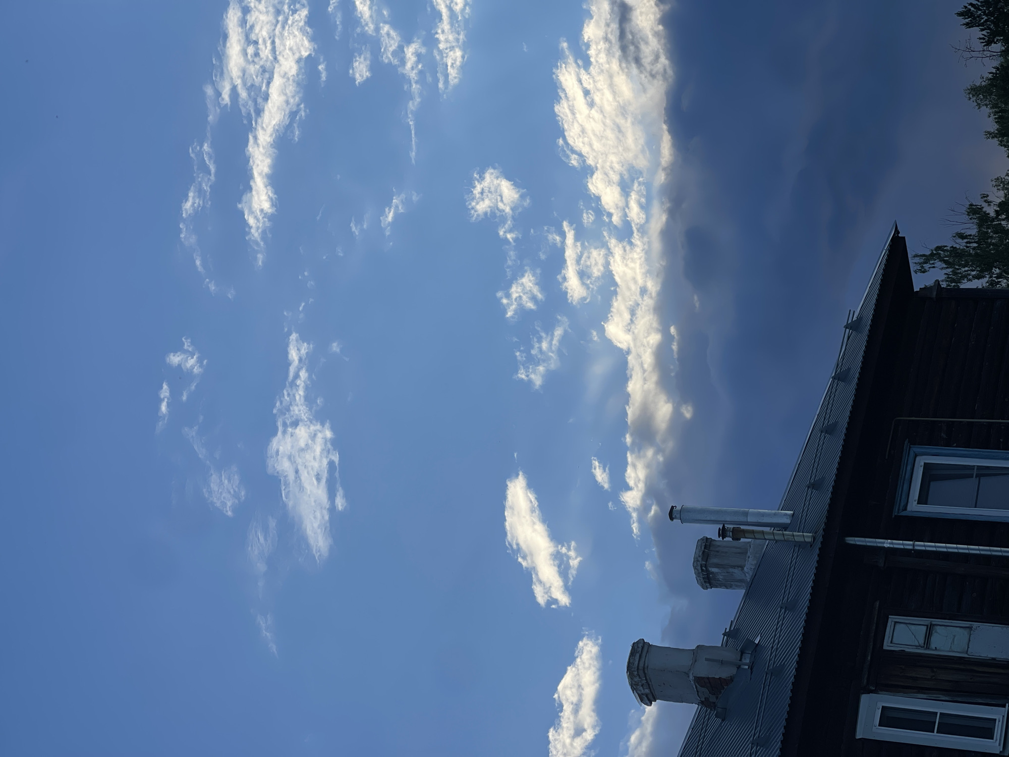 The image shows the roofline of a dark wooden building with a corrugated metal roof against a bright blue sky filled with scattered white and grey clouds. Three chimneys are visible on the roof: the leftmost is a wide, aged brick chimney; the middle chimney is a narrower, older brick structure; and the rightmost is a tall, silver metal chimney with a circular base. The building has several windows with white frames, some appearing weathered. In the distance, the tops of green trees are visible below the right side of the building. The scene appears to be set in a rural or suburban area, possibly on the outskirts of the city of Заречный, Russia, given the architectural style and the presence of natural elements. The lighting suggests it is daytime, possibly late afternoon or early evening due to the varied cloud cover and potential for approaching weather.