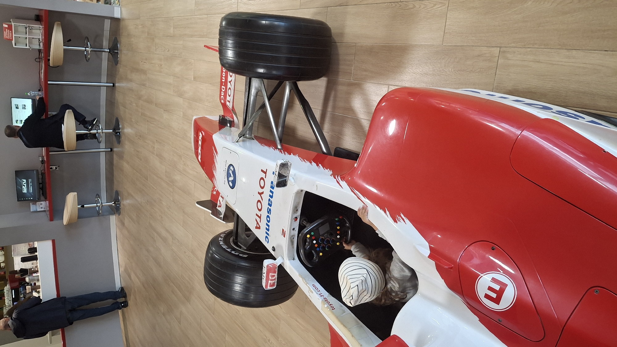 The image depicts the interior of a public space with a red and white Formula 1 racing car as the central subject. The car's cockpit is occupied by a child wearing a striped hat, who is holding the steering wheel. The car has "TOYOTA" and "Panasonic" branding. In the background, two men are seated at a red counter with computers. One man is facing a monitor, while the other is looking away from the camera. There are bar stools around the counter. The flooring is made of light-colored wood planks. The location is identified as Novogorelovo, Russia.