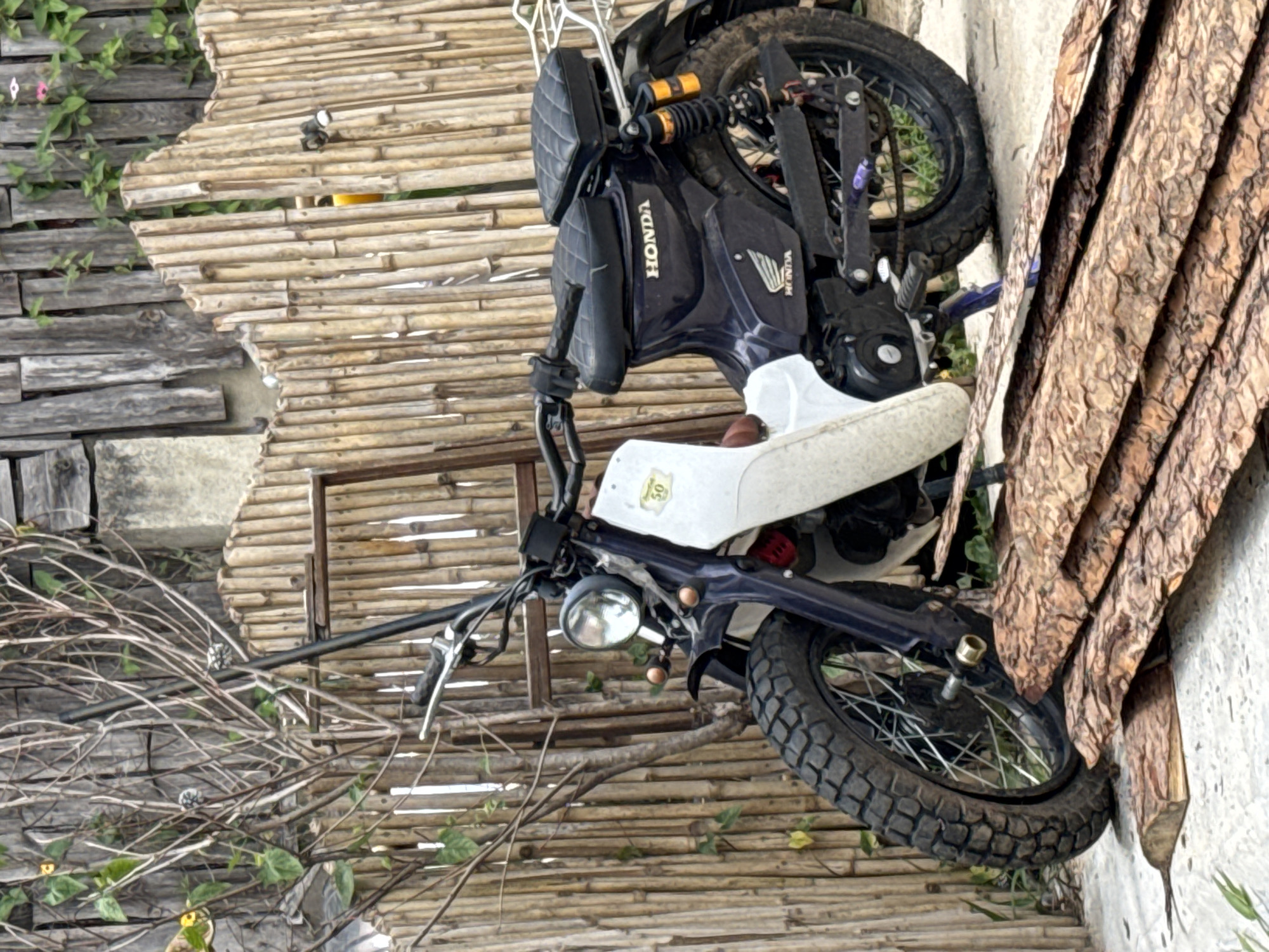 A motorcycle with a white seat is leaning on a wall covered with bark strips. The motorcycle is parked on a bamboo surface. The word "HONDA" is visible on the side of the motorcycle. Branches and vines are visible in the foreground. The location appears to be outdoors.