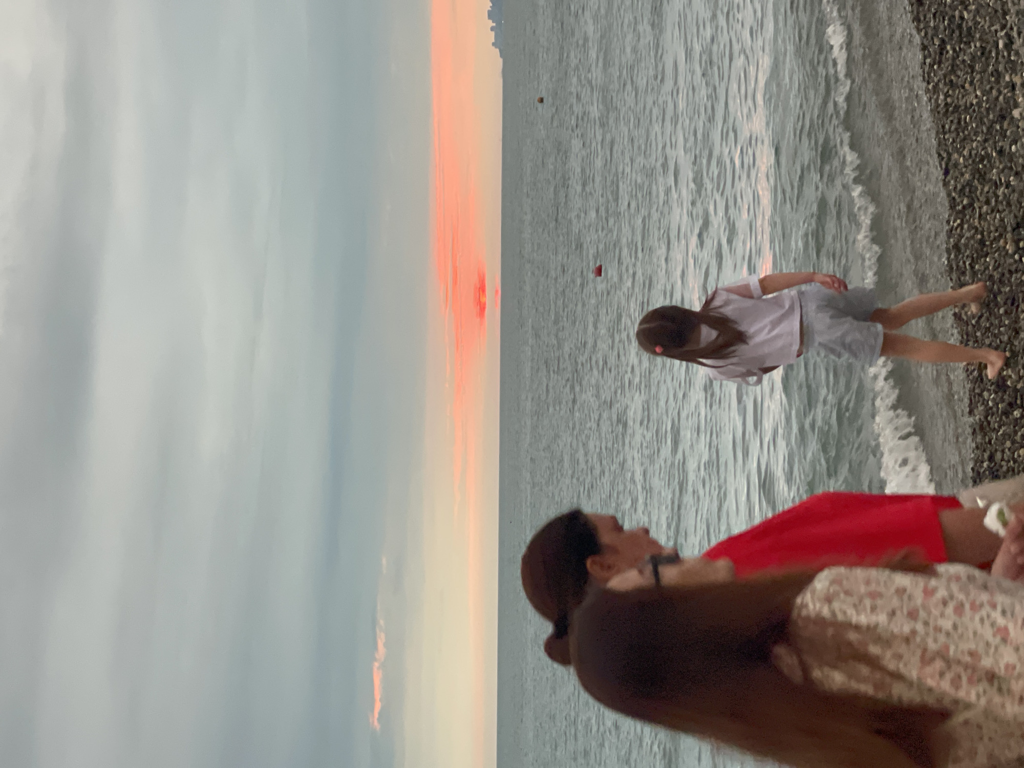 The image depicts a sunset over the Black Sea in Sochi, Russia. Three people are visible on the pebble beach. In the foreground, two women are walking from left to right, their backs partially to the camera. The woman on the left has long, dark hair and is wearing a floral patterned top. The woman next to her has her hair pulled back in a bun and is wearing a bright red top. Further down the beach, a young girl with her back to the camera is standing by the water's edge. She has dark hair tied in a ponytail with a pink bow and is wearing a white t-shirt and light-colored shorts. The sea is calm with small waves lapping the shore. In the distance, the setting sun casts a vibrant orange and pink glow across the horizon, illuminating the clouds. A city skyline is faintly visible on the far right horizon. Two buoys, one orange and one red, are visible in the water. The sky is partly cloudy with shades of grey and pink.