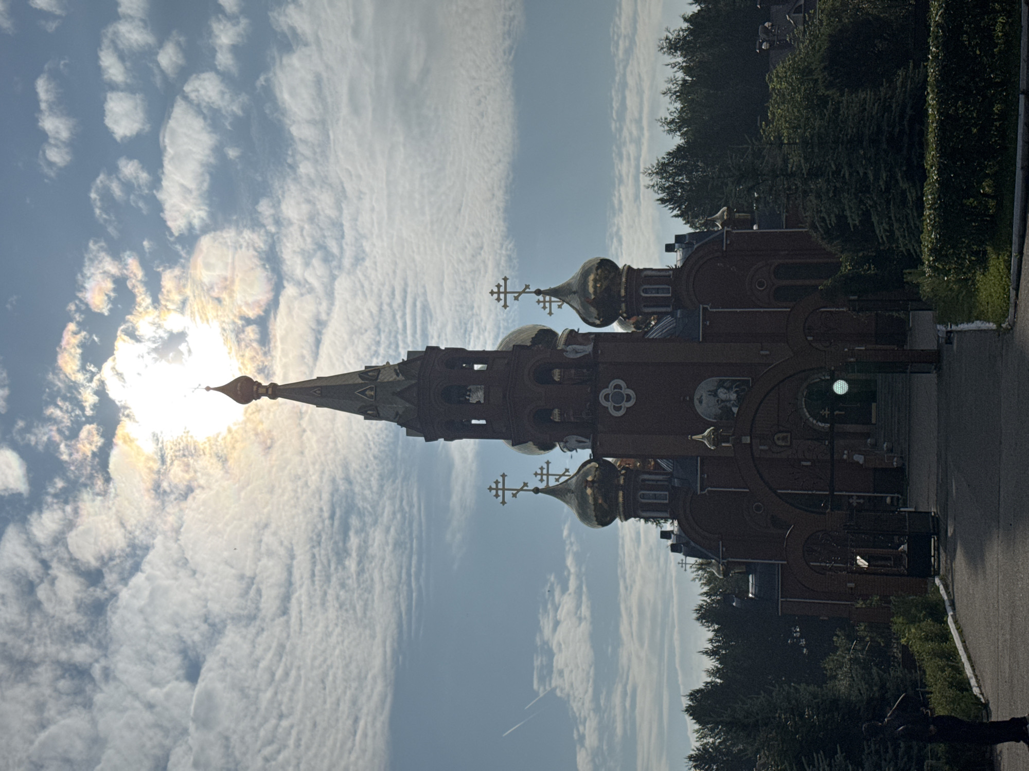 An Orthodox church with red brick walls and golden domes stands prominently against a cloudy sky, with the sun partially obscured by clouds, creating a lens flare effect. The church features multiple crosses on its domes and a tall, spired bell tower. In the foreground, a paved walkway leads to the church entrance, which is framed by an arched gateway. Lush green trees and manicured bushes surround the church grounds. To the left of the walkway, a dark silhouette of a person stands with their hands behind their back. The image appears to be taken during the daytime, possibly in the morning or late afternoon, judging by the quality of light and the direction of the sun. The location is the Church of the Holy Trinity in Chaikovsky, Russia.