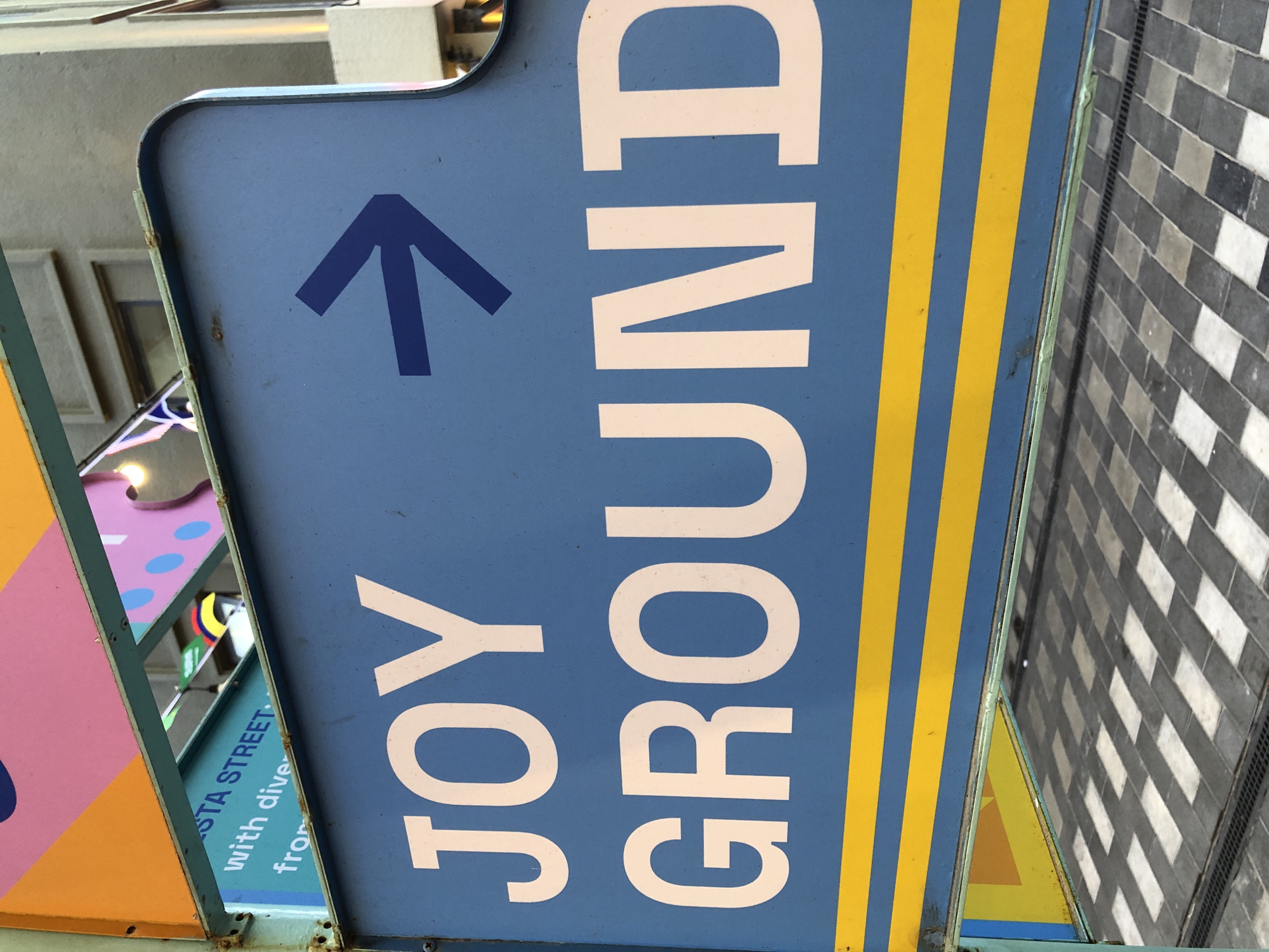 A blue directional sign with white text reads "JOY GROUND" and has a blue arrow pointing to the right. The sign is positioned in an urban environment, potentially in Đà Nẵng, Vietnam, with colorful panels above and a patterned tiled surface below. The overall scene suggests a public space or an entertainment venue.