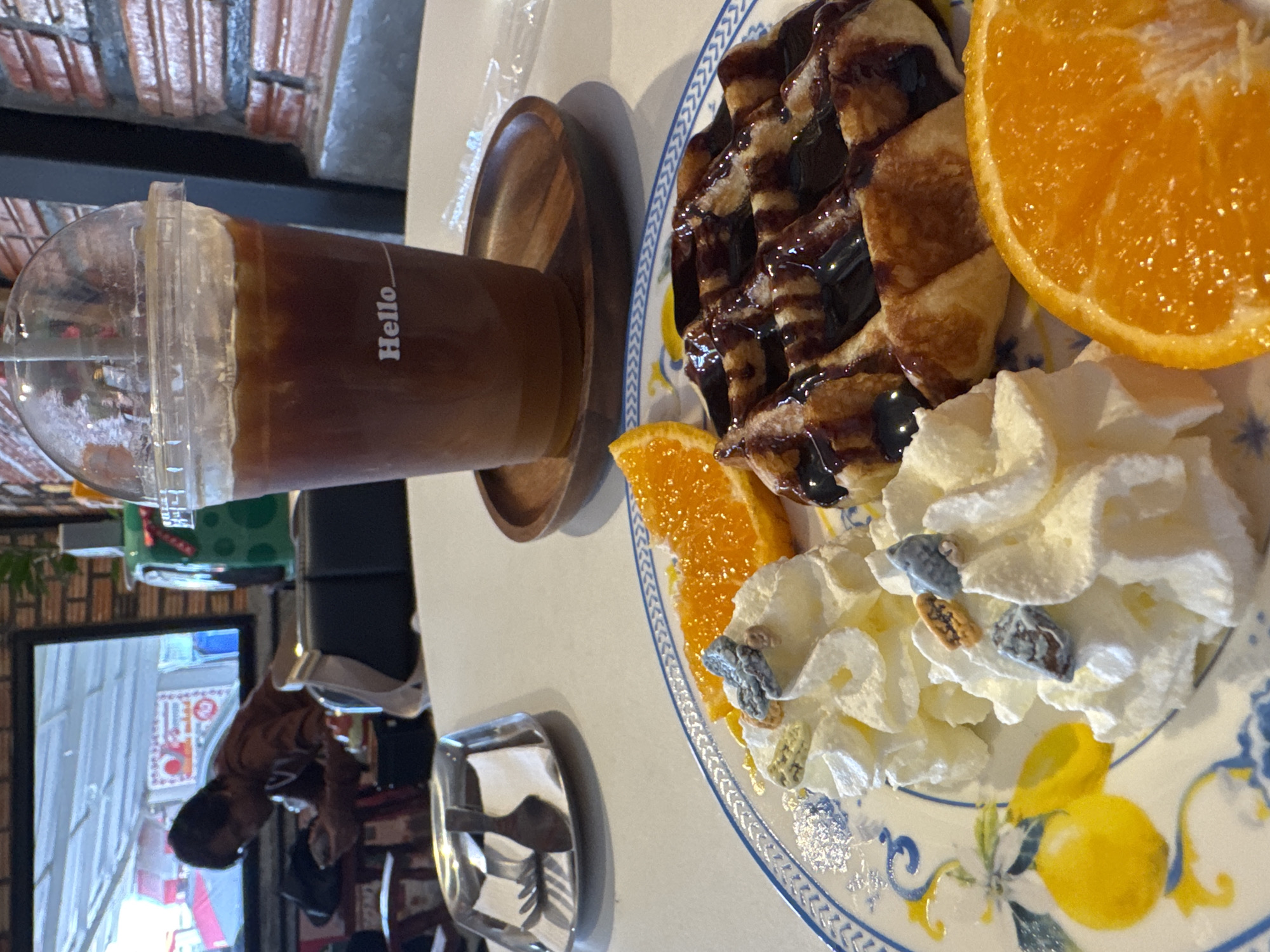 A close-up, eye-level shot captures a table setting in a brightly lit cafe. In the foreground, a plate displays a Belgian waffle drizzled with chocolate sauce, two dollops of whipped cream topped with colorful sprinkles, and a slice of orange. To the right of the plate is a clear plastic cup filled with iced coffee, bearing the "Hello" logo, resting on a wooden coaster. In the background, a person is visible through a television screen, and a brick wall is to the right. The environment suggests a cafe located in Surat Thani City Municipality, Thailand.