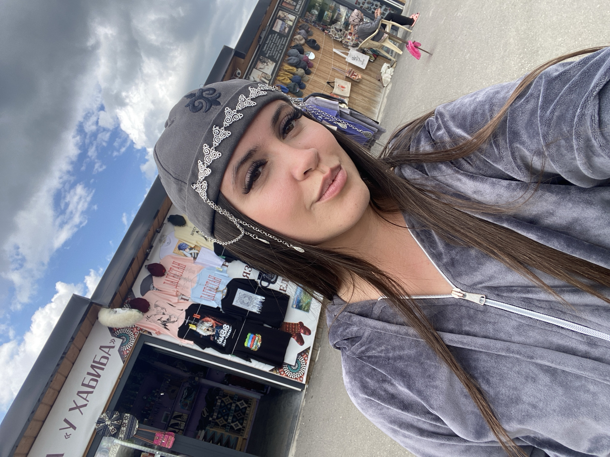 A young woman with long, dark hair and noticeable eyelashes is taking a selfie outdoors. She is wearing a grey velour hoodie and a grey skullcap adorned with silver detailing and chains. She is standing in front of a market stall with various items displayed, including t-shirts with graphics, hats, and bags. The stall appears to be in an open-air market setting, with other stalls and people visible in the background. The sky is partly cloudy. The location is the city of Tsalkita, Russia.