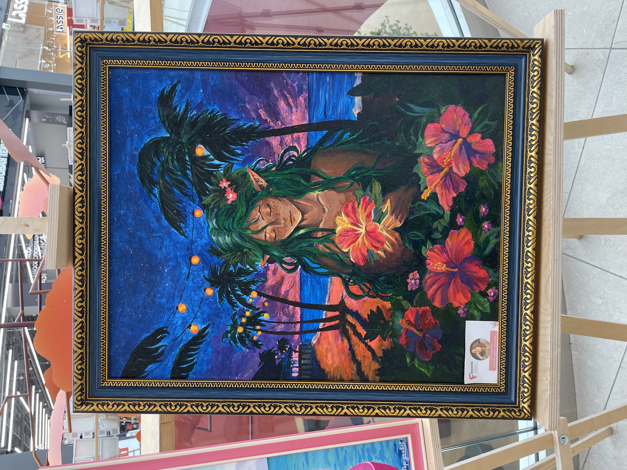 A framed, colorful acrylic painting of a mythical creature with green hair and pointed ears sits on an easel. The creature, likely a nature spirit or elf, is depicted holding a bright pink flower amidst lush foliage, with a tropical beach scene featuring palm trees, string lights, and a starlit sky in the background. The painting is displayed at an exhibition in Perm, Russia, as indicated by a business card with Russian text and a photo of the artist, Evgenia Nekrasova, affixed to the bottom of the frame. Other artworks and display structures are visible in the blurred background, suggesting an indoor art fair or gallery setting.