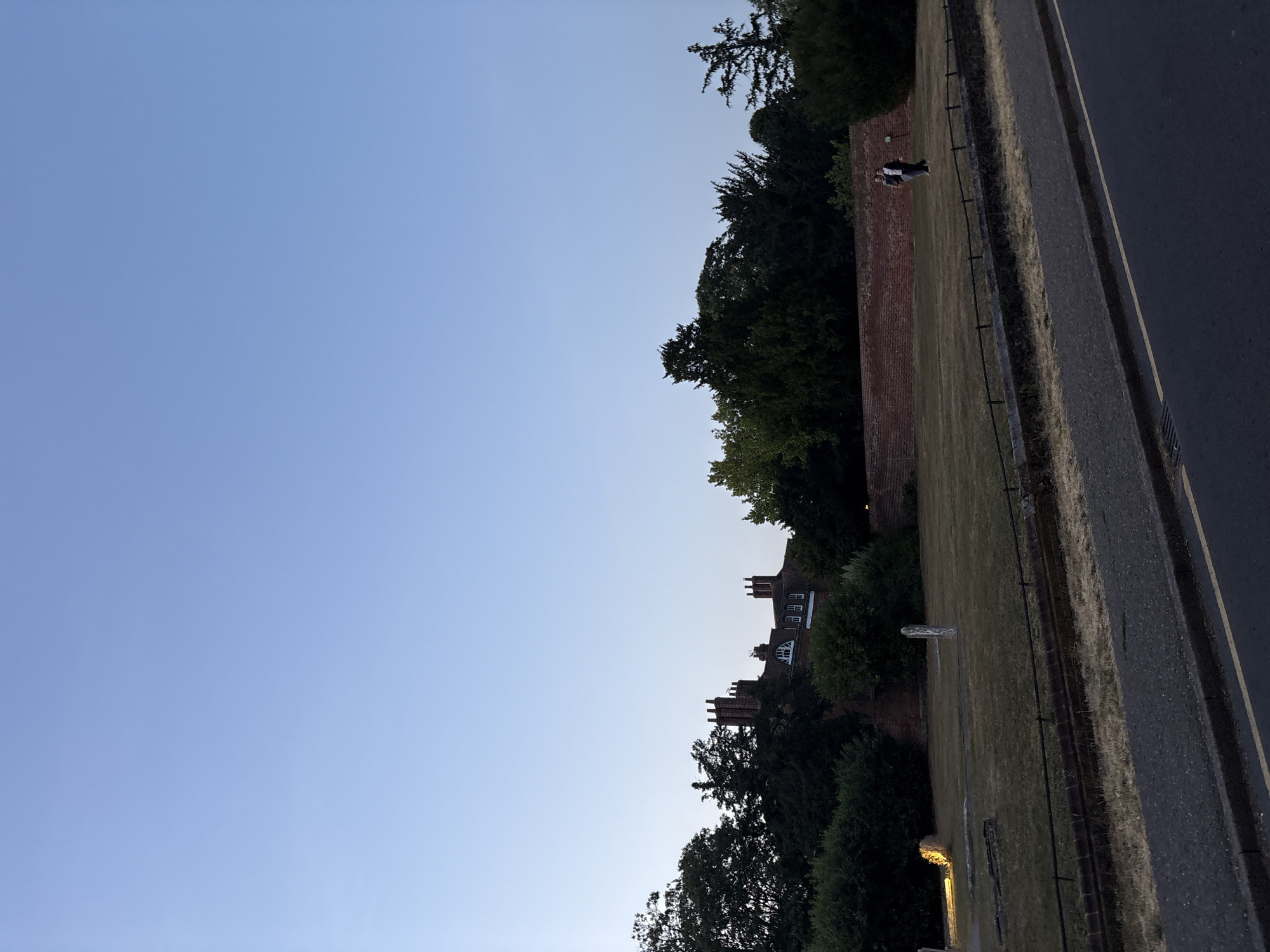 This is a photograph of the Essex Regiment Museum and Gardens, located in Chelmsford, United Kingdom. The image captures a wide view of the gardens with the museum building visible in the background.
In the foreground, a paved road runs along the bottom of the frame, with a sidewalk and a low brick wall separating it from the garden. The garden itself is covered in dry grass with several stone markers and a single illuminated object on the left. A black metal fence runs through the garden.
In the middle ground, a red brick wall stretches across the scene, separating the garden from the area behind it. Two people are walking on the grass in front of the wall. Behind the wall, the museum building is visible, characterized by its red brick facade and multiple chimneys. A line of trees forms a dense green backdrop behind the building and stretches across the horizon.
The sky is clear and a pale blue, suggesting it is either early morning or late evening. The overall lighting indicates it is dusk or dawn. The scene is serene and quiet, with no other discernible activity.