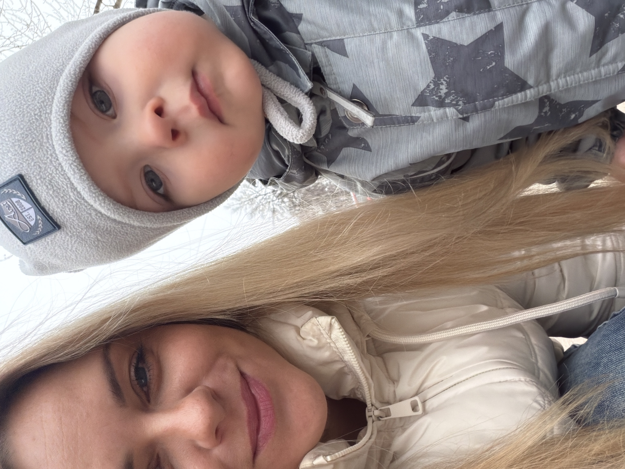 A selfie taken outdoors in daylight features a woman and a baby. The woman has fair skin, blond hair, and is wearing a white puffer jacket and blue jeans. The baby, who appears to be lying down above the woman, has fair skin, a grey hat with a patch, and a grey jacket with dark grey stars. The environment appears to be wintery with white or light-colored blurred background suggesting snow or ice. Given the location constraint, the image was likely taken in Votkinsk, Russia.